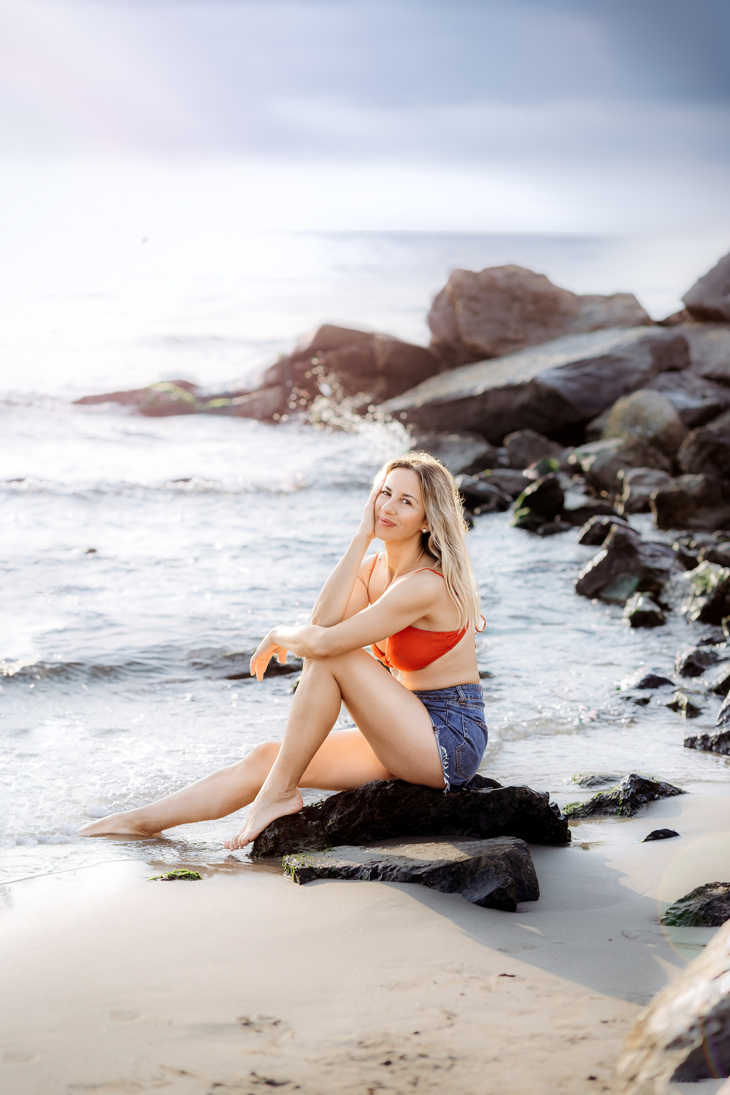 Улыбающаяся женщина в красном топе от бикини и джинсовых шортах сидит на прибрежных скалах у воды во время золотого часа в Кальпе, Испания — яркая и естественная пляжная портретная фотография, передающая радость, мягкий свет и красоту Средиземноморья, идеально подходит для портретных фотосессий в Кальпе и Испании.