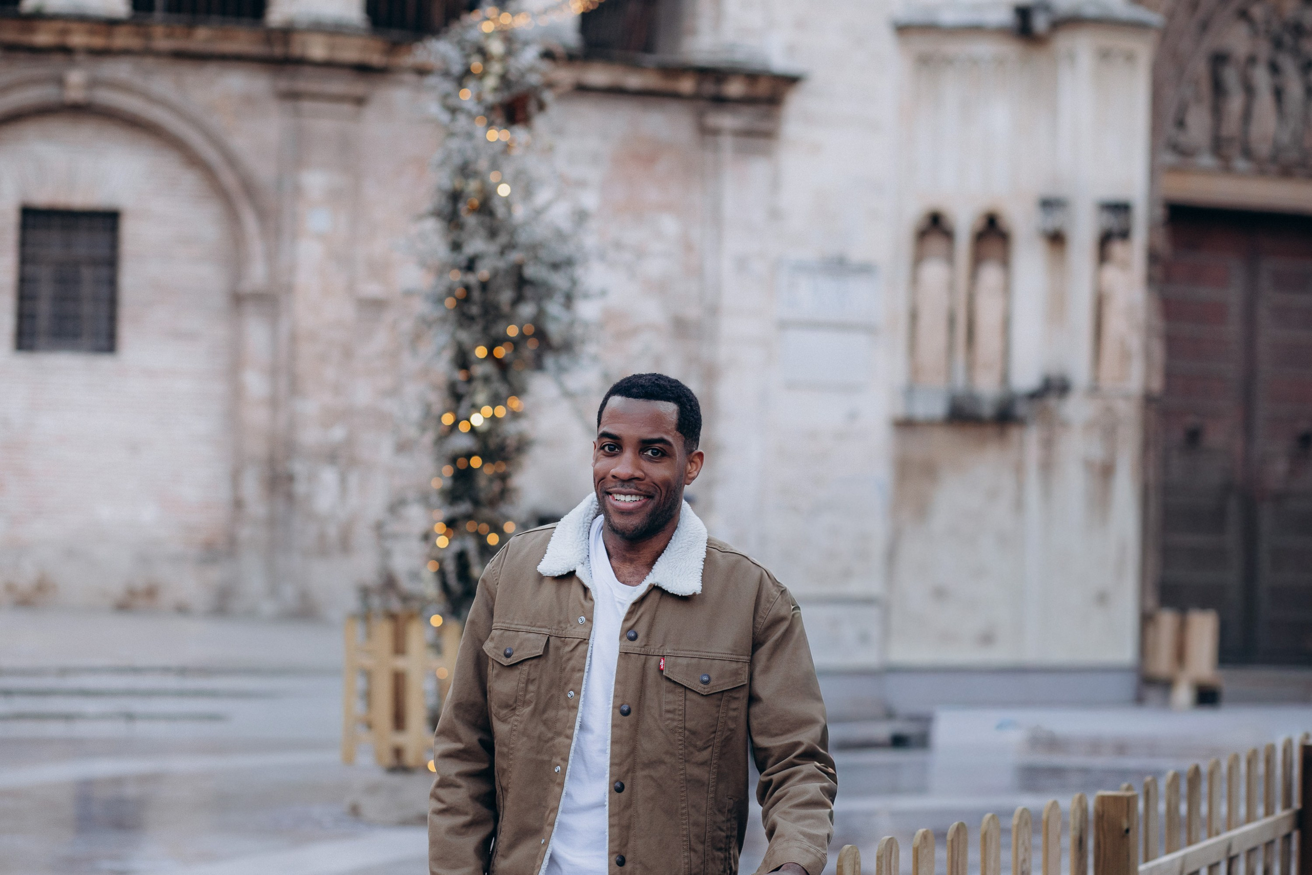 Мужская портретная фотосессия в Валенсии, Испания, с улыбающимся мужчиной в повседневной куртке, позирующим в исторической городской локации с мягким естественным светом и праздничными боке-деталями на фоне, создающими тёплое и аутентичное городское настроение — идеально для мужской портретной фотографии, lifestyle-фотосессий для мужчин, городских портретов при естественном освещении, изображений для персонального бренда и профессиональных мужских фотосессий в Валенсии и по всей Испании.