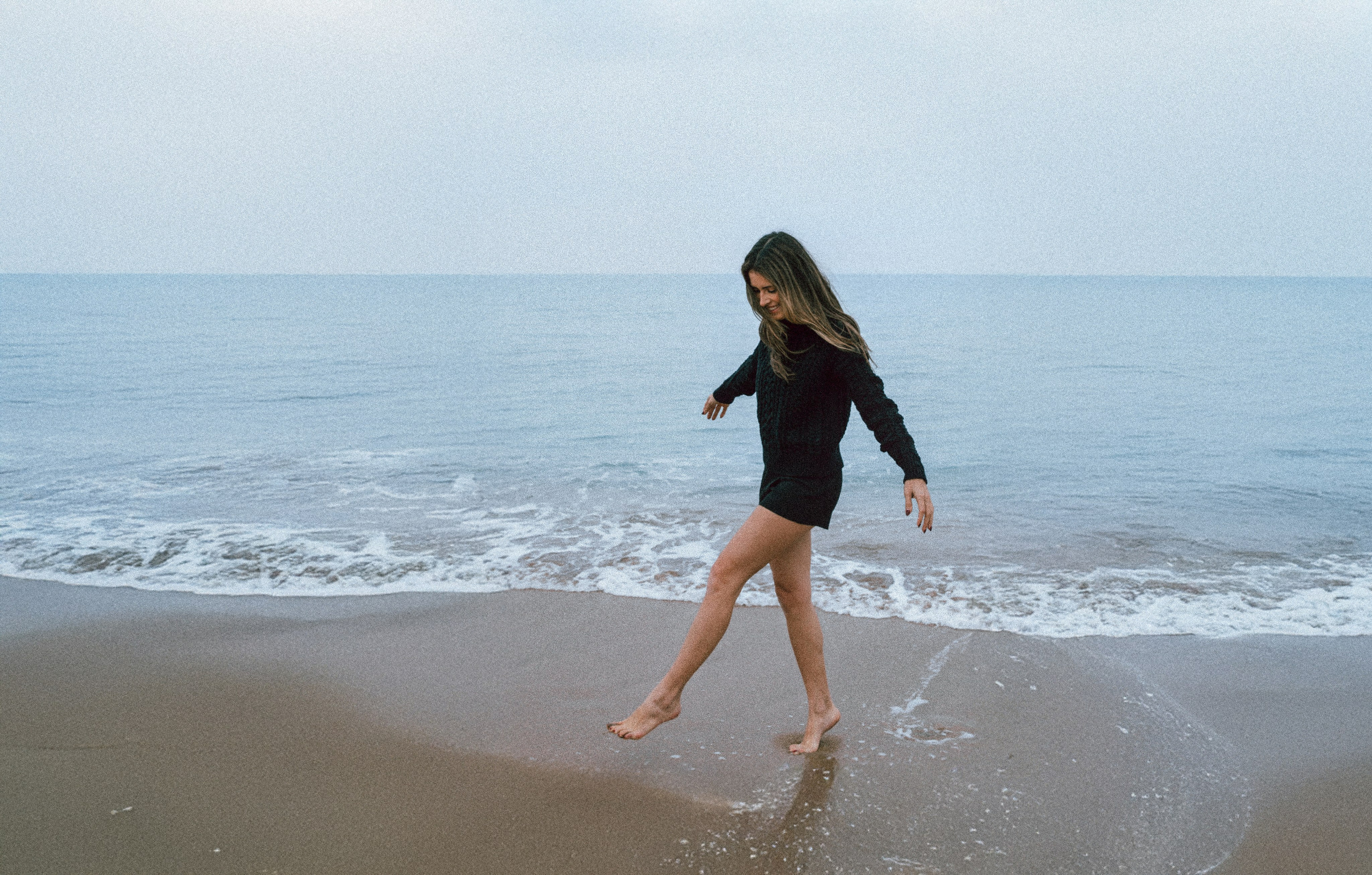 Повседневный пляжный портрет женщины, идущей вдоль берега босиком в черном свитере и шортах, с волнами, нежно касающимися ее ног, в Валенсии, Испания