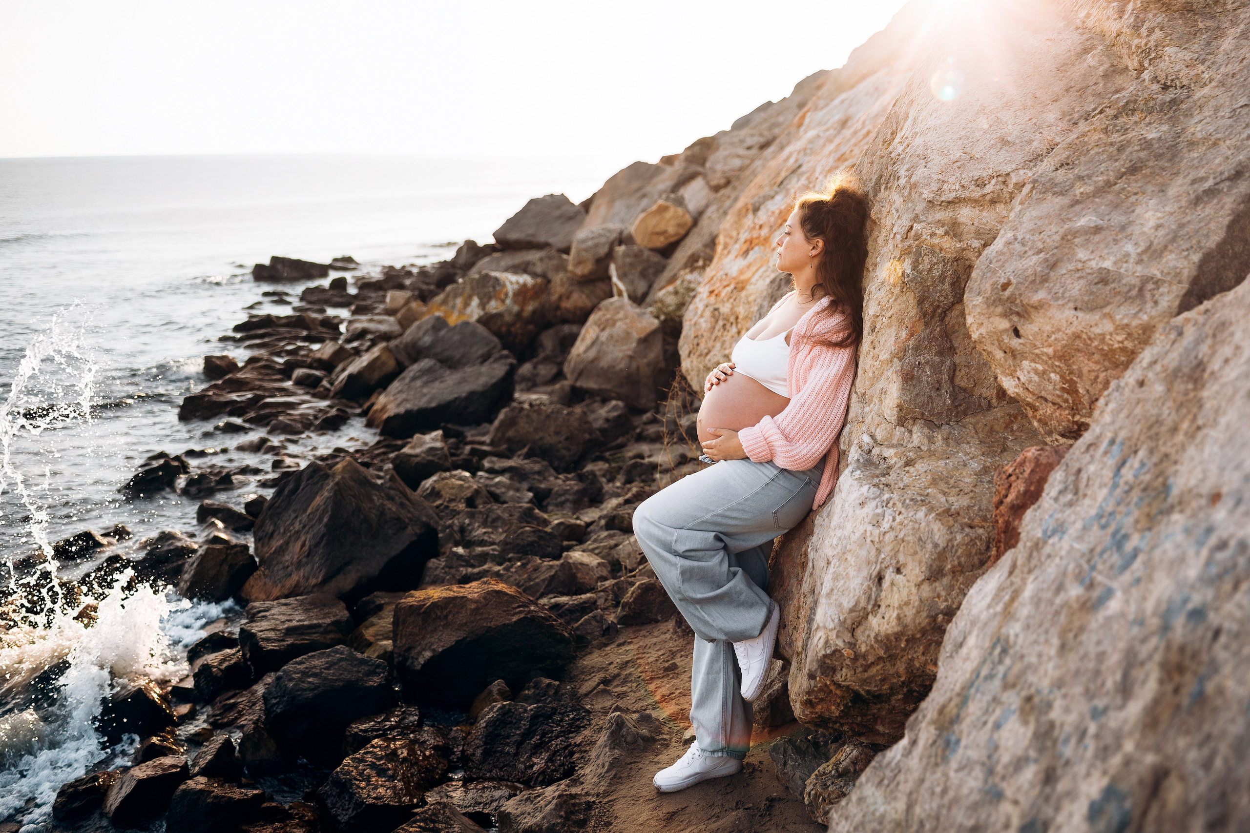 Sesión de fotos de embarazo en Valencia, España, con una mujer embarazada posando sobre rocas costeras junto al mar, capturada con cálida luz natural para una tranquila y cinematográfica escena de embarazo en la playa — ideal para sesiones maternity en Valencia y España.