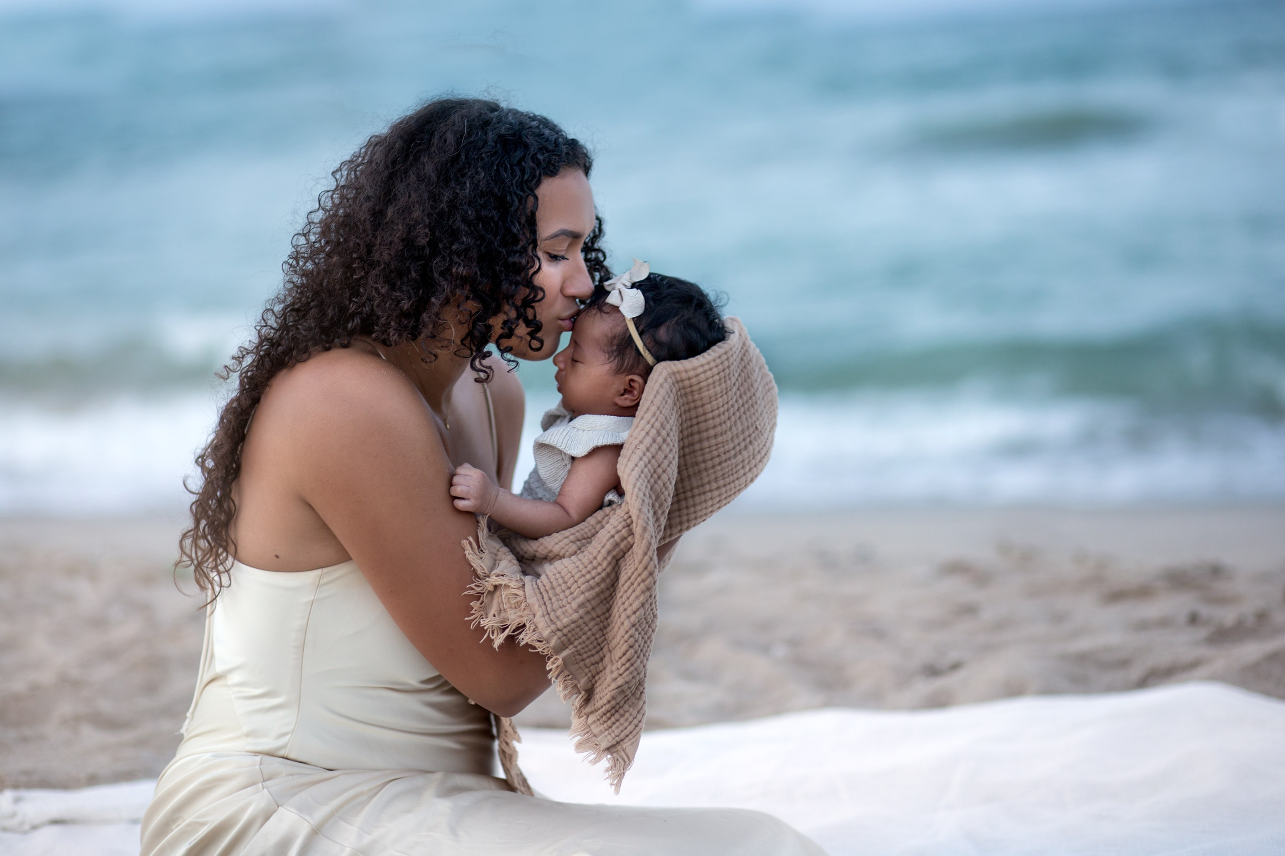 Retrato tierno en la orilla de la playa de una madre besando la frente de su bebé envuelto en una manta suave — un momento emotivo capturado en la arena de Valencia, España.