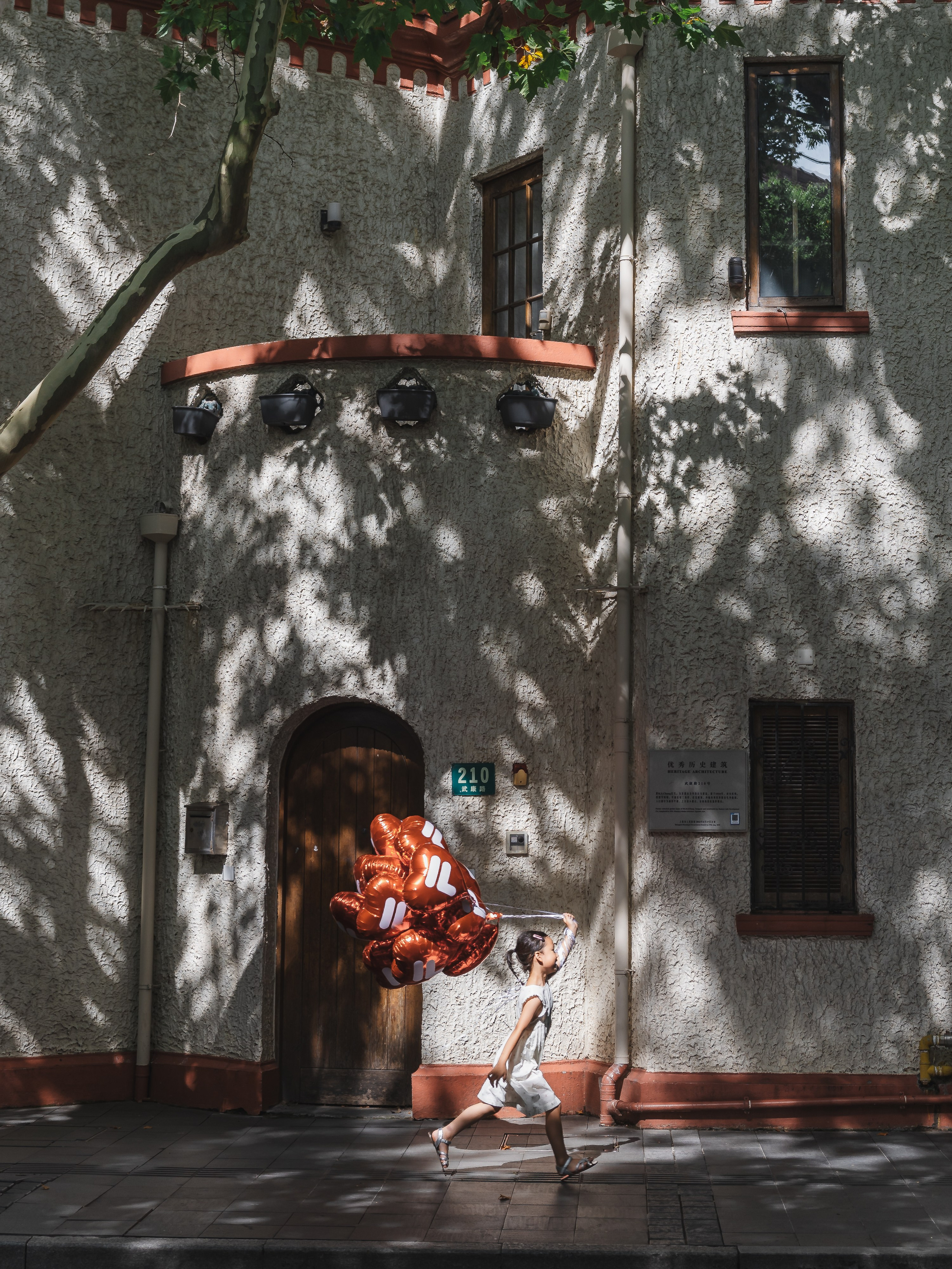 In the dancing light and shadows, a five-year-old girl holds a balloon and runs past an old house on a street lined with sycamore trees in Shanghai.