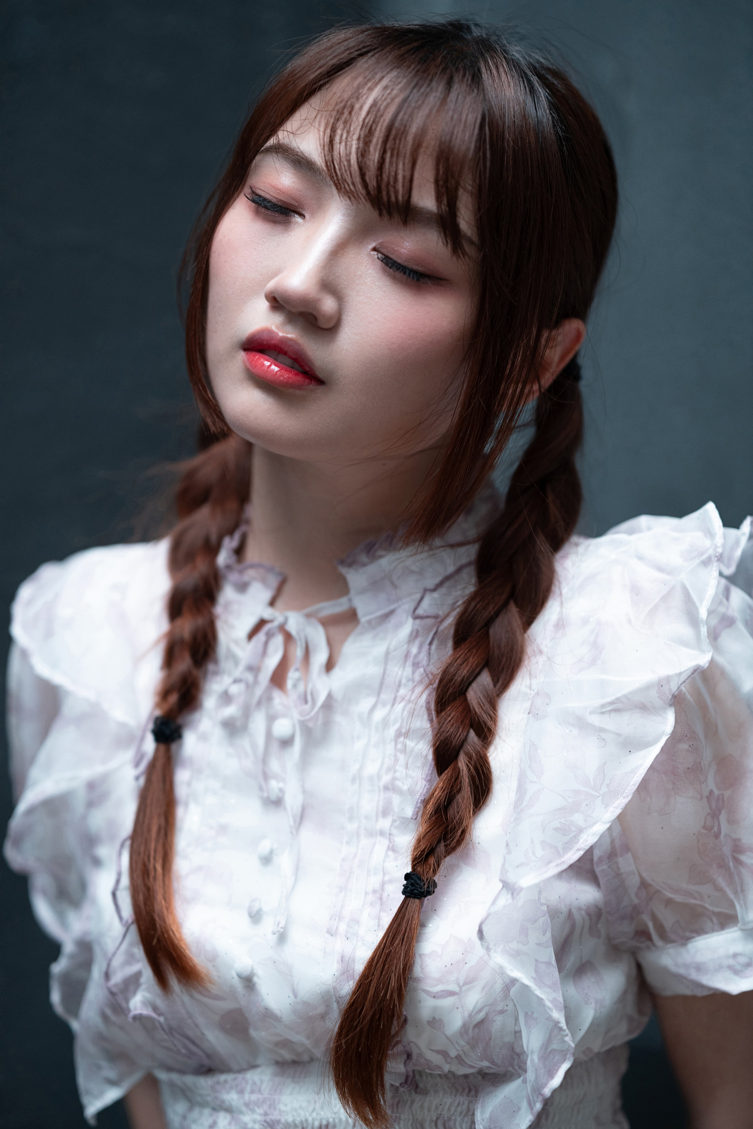 Close-up portrait of the model with eyes closed, peach-pink makeup and twin braids, softly lit against a dark teal background.