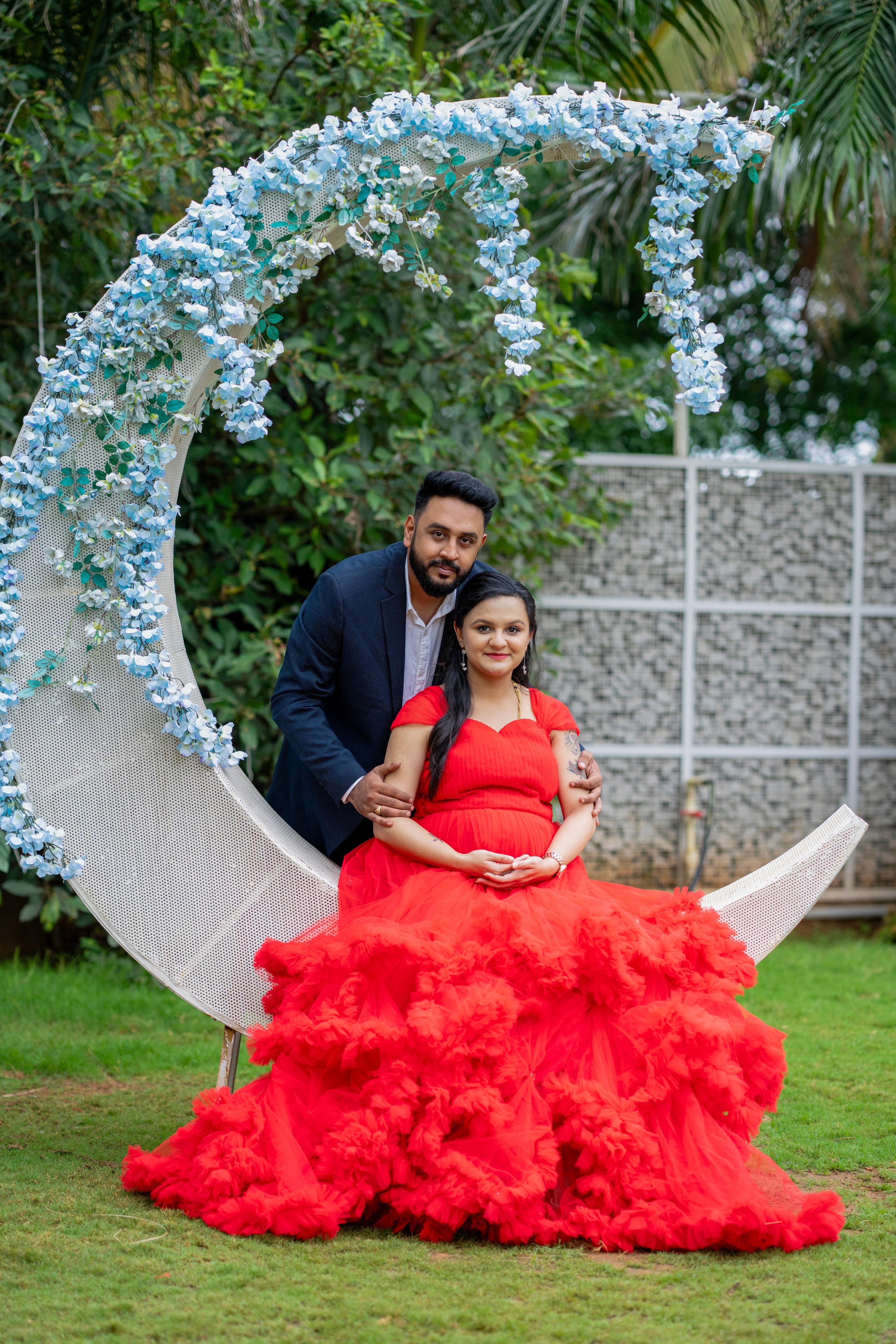 Outdoor maternity photoshoot in Bengaluru featuring a man in a navy suit and a woman in a red ruffled gown sitting on a crescent moon prop with blue flowers.