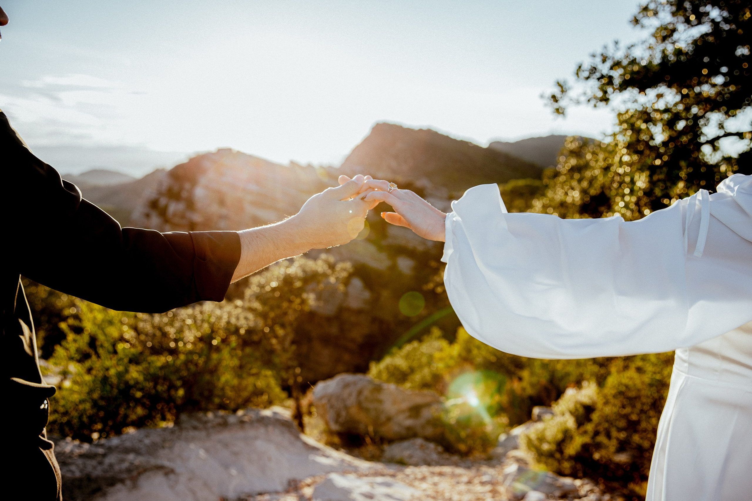 Engagement photoshoot in València, Spain, capturing an intimate close-up of a couple holding hands against a sunlit mountain landscape and Mediterranean greenery — a romantic, symbolic wedding love story image ideal for engagement photography, pre-wedding sessions, elopements, and professional wedding photoshoots in València and across Spain.