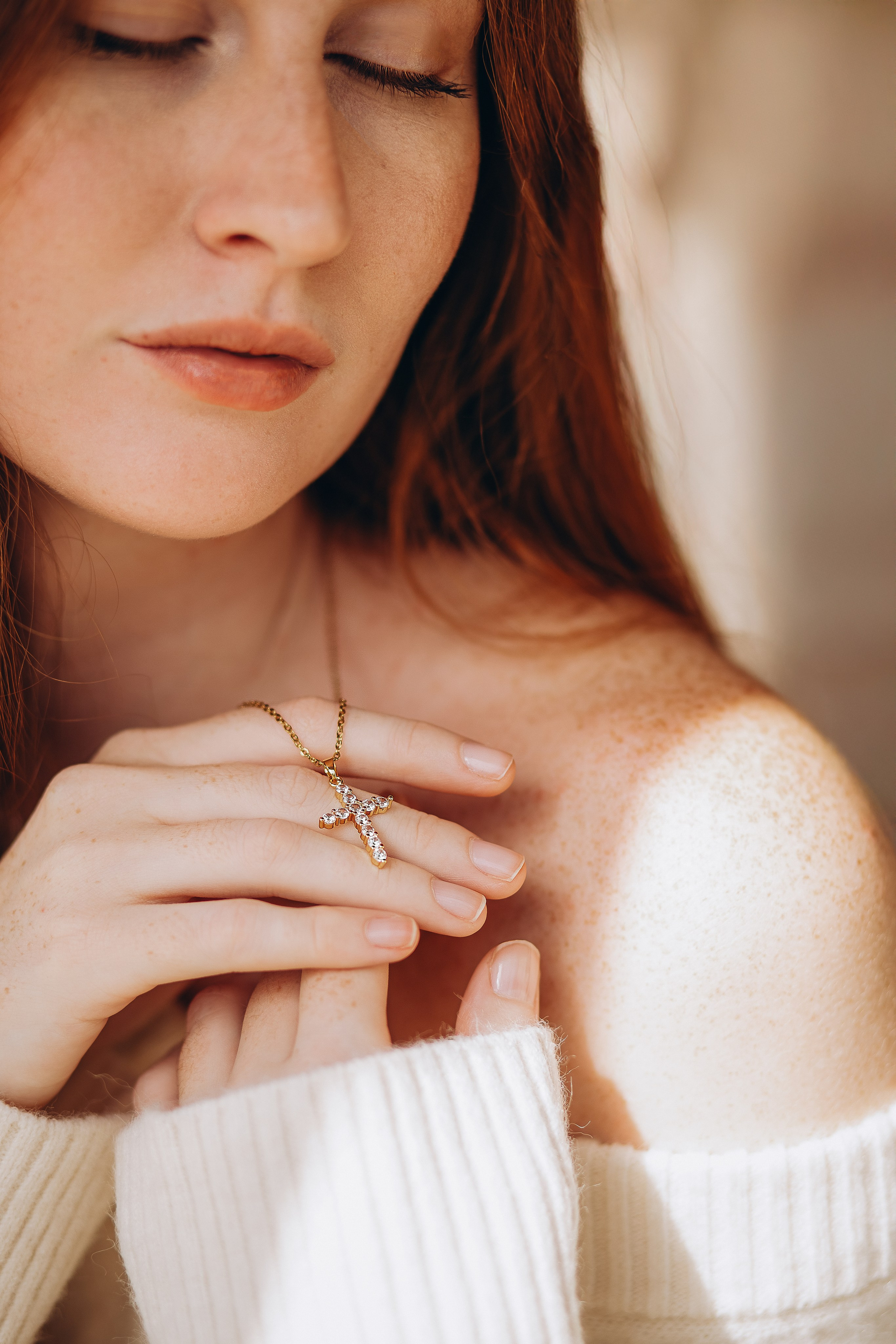 Close-up brand photo of a woman with closed eyes gently holding a gold cross necklace, highlighting minimalist beauty and fine jewelry. Captured in Valencia, Spain — perfect for showcasing delicate product details and emotional branding in lifestyle content and product photoshoots.