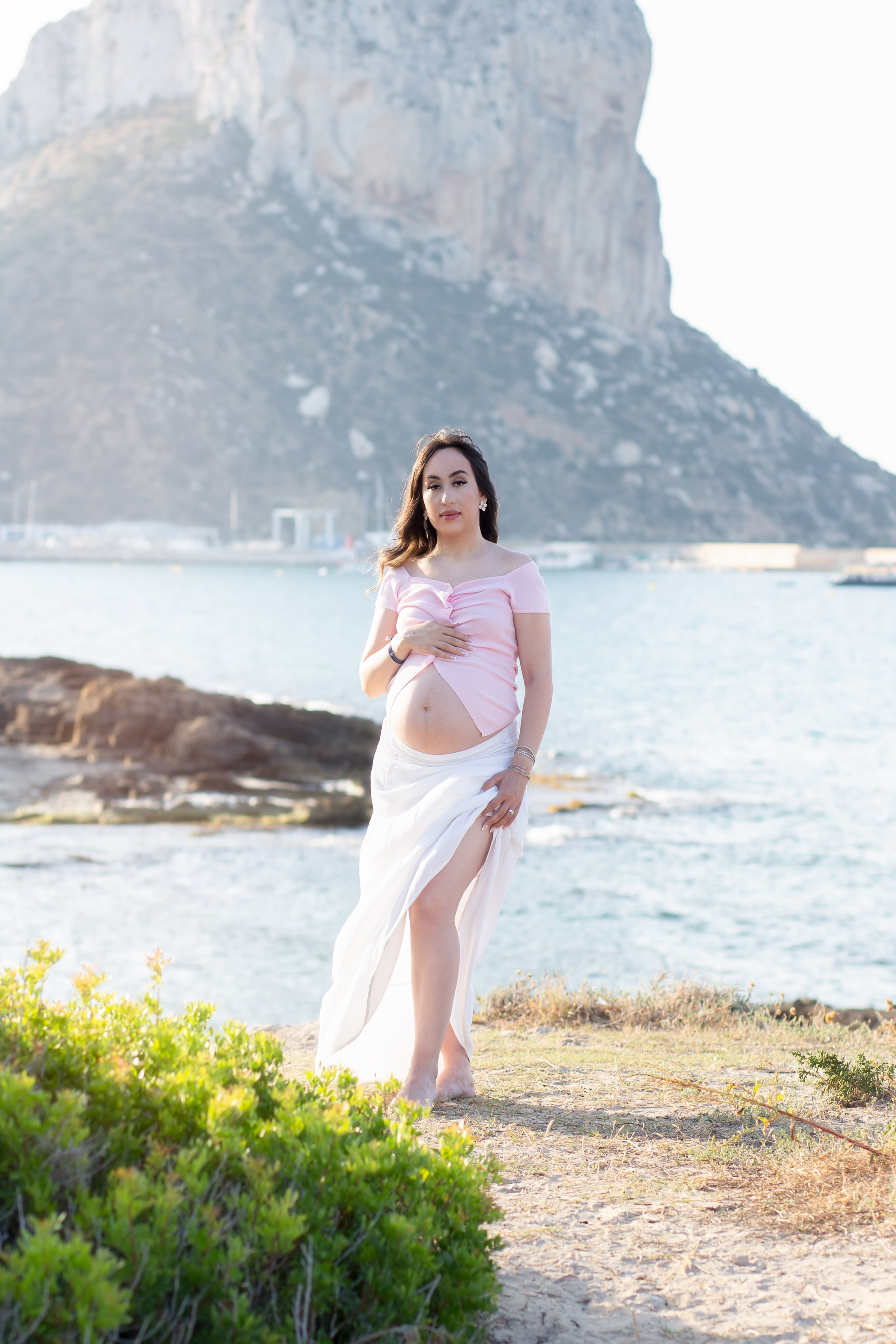 Elegante sesión de fotos de maternidad en Calpe, España — mujer embarazada con falda blanca fluida y top rosa, descalza junto al mar con el Peñón de Ifach al fondo. Una sesión playera de ensueño y emocional ideal para fotos de embarazo en Calpe y la costa española.