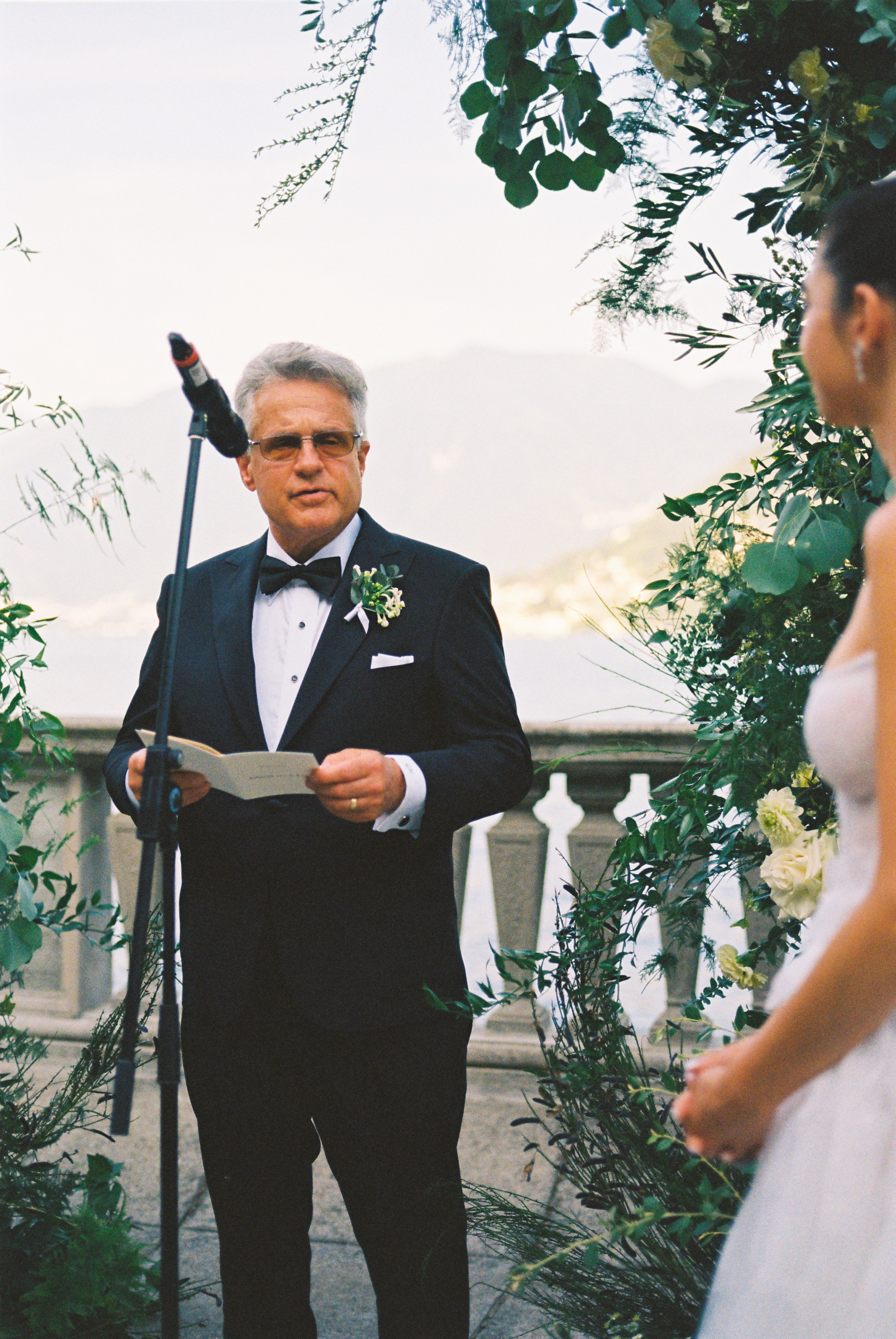 Officiant in tuxedo delivers a heartfelt speech at wedding ceremony, with scenic lake backdrop.