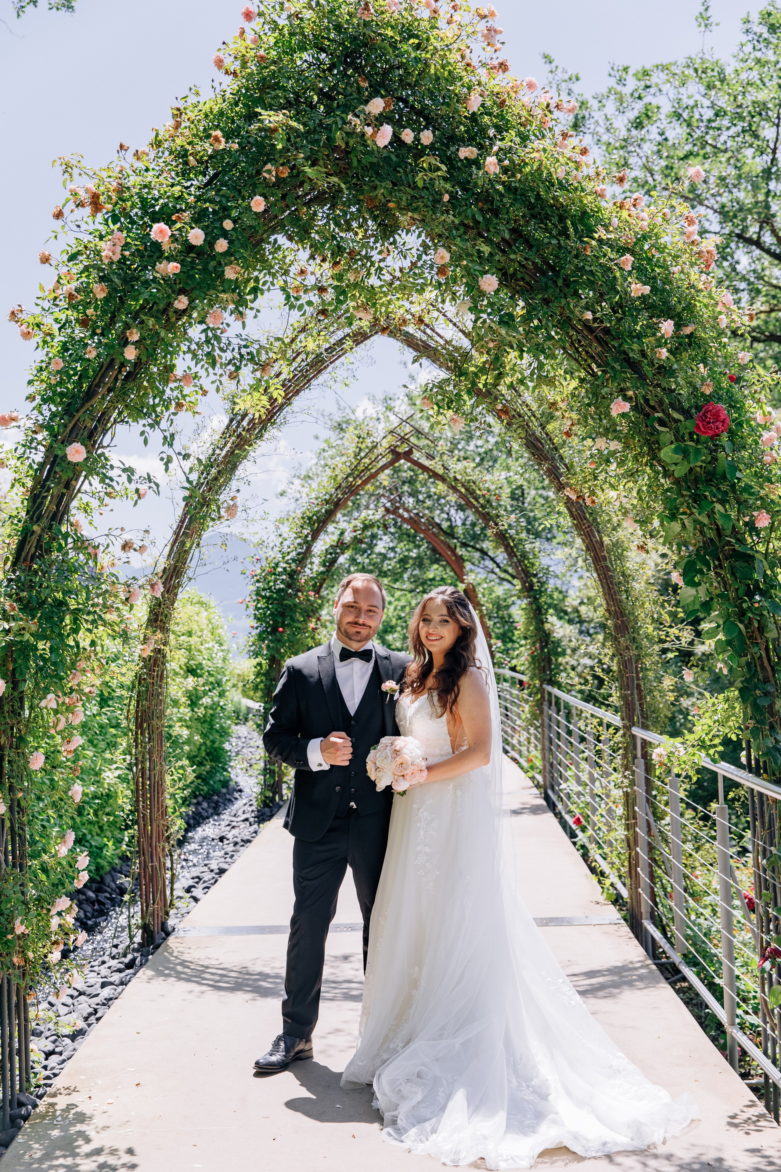 Der Bräutigam und die Braut stehen unter einer Rosenbogen im Garten von Schloss Trauttmansdorff, umgeben von einer malerischen Gartenlandschaft.