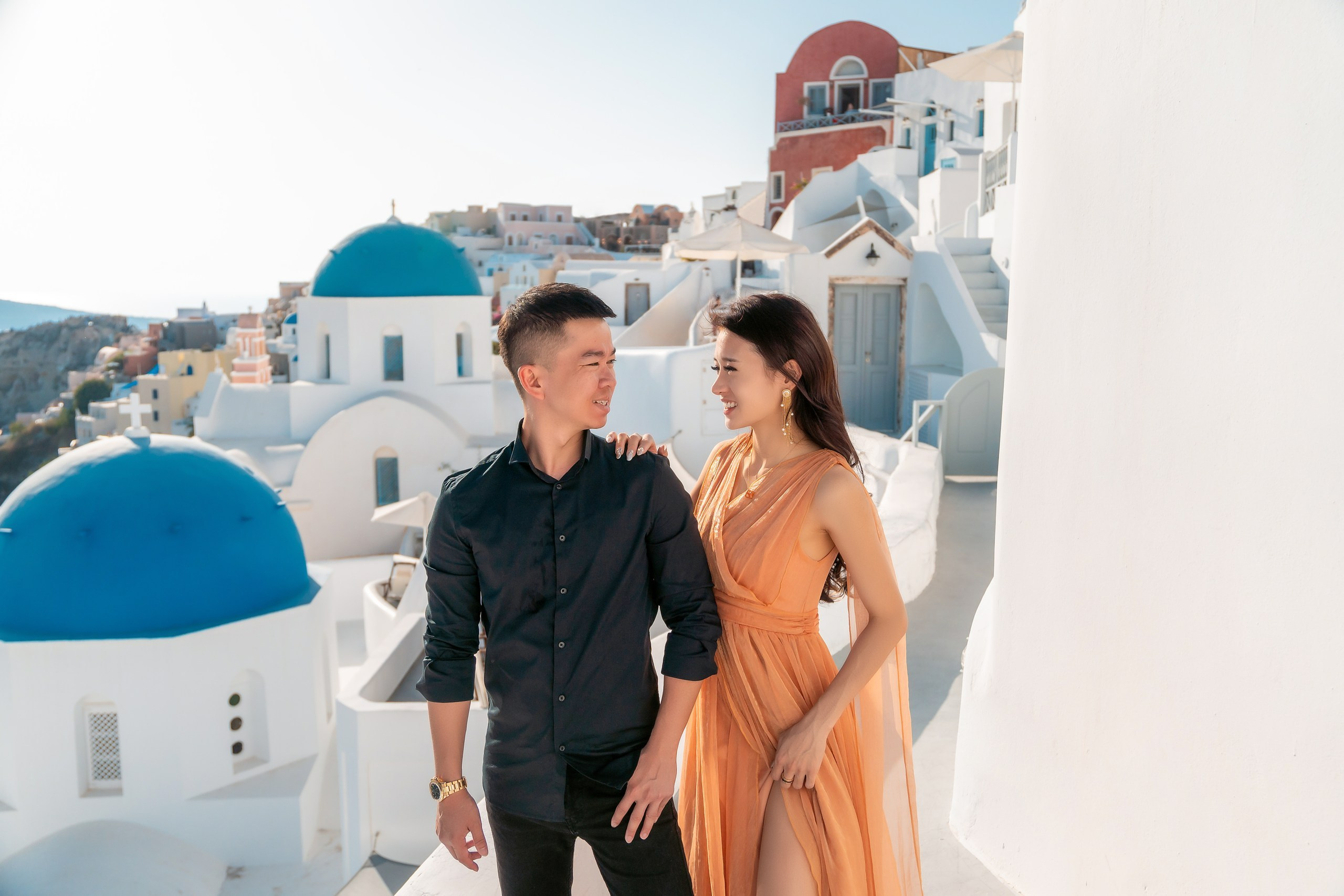 A romantic photoshoot in Oia, Santorini, featuring a couple posing against the iconic whitewashed buildings and blue-domed churches. The man stands confidently in a black shirt, while the woman glows in a flowing orange dress, with the stunning Aegean Sea backdrop enhancing the picturesque scene.