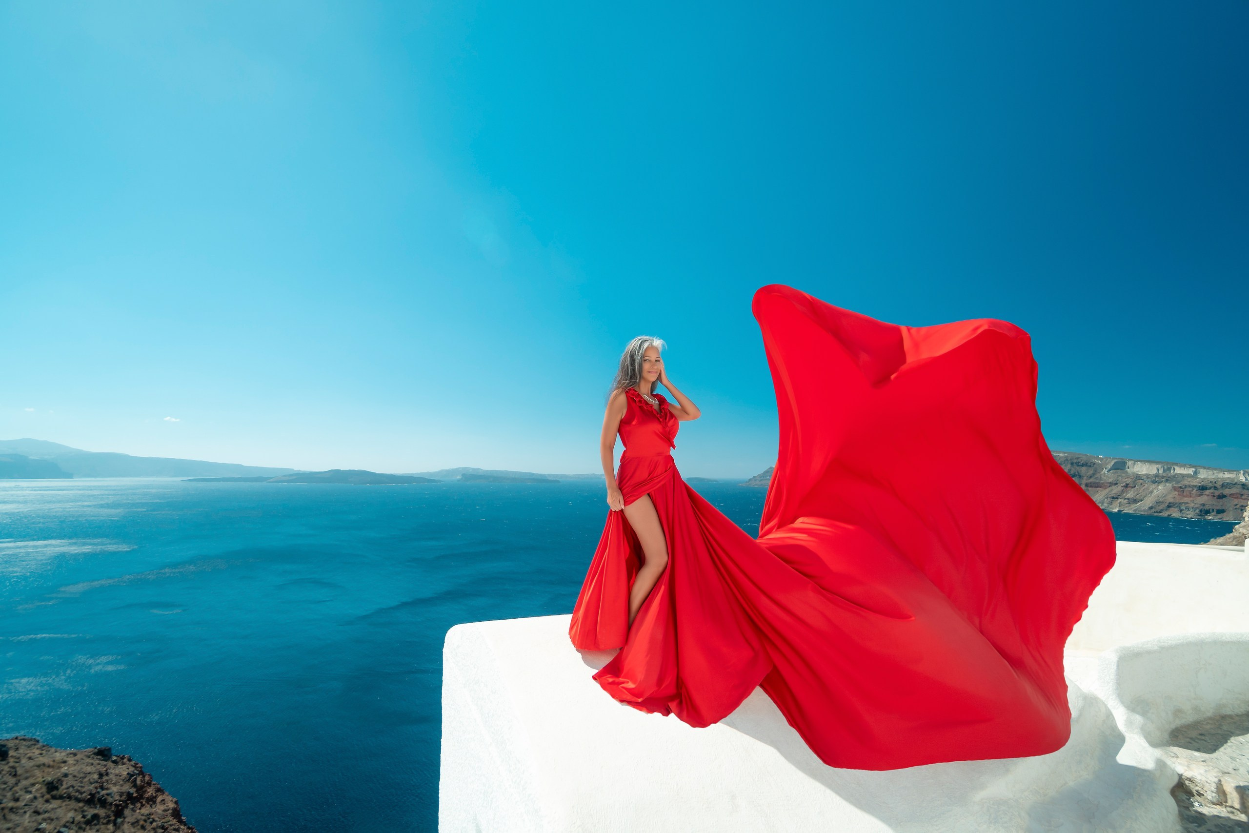 Red plus size flying dress with ruffles and V-shaped back, flowing dramatically against Santorini's blue-domed cliffs.