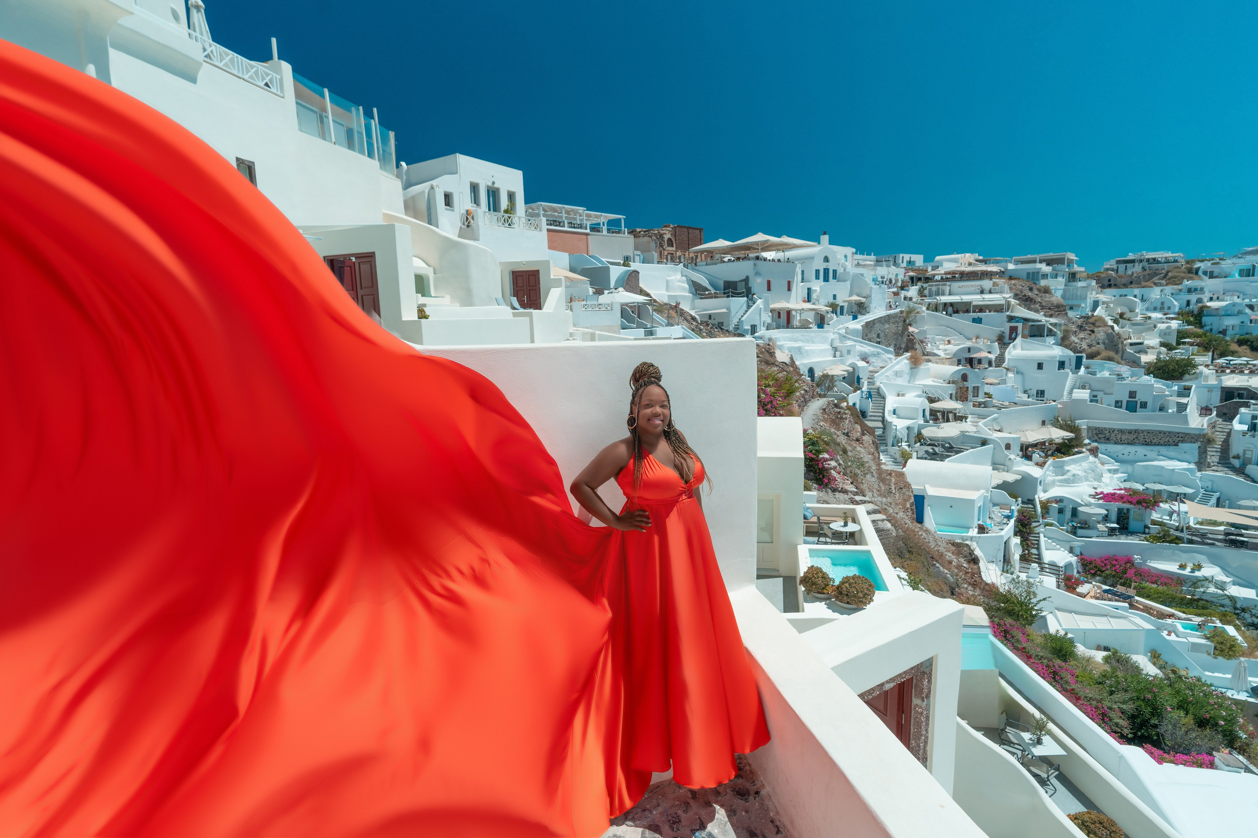 "Rent this striking orange-red flying dress for your Santorini photoshoot, flattering for plus sizes. Sokolove’s photography showcases its elegance and dynamic flow for a stunning visual impact."