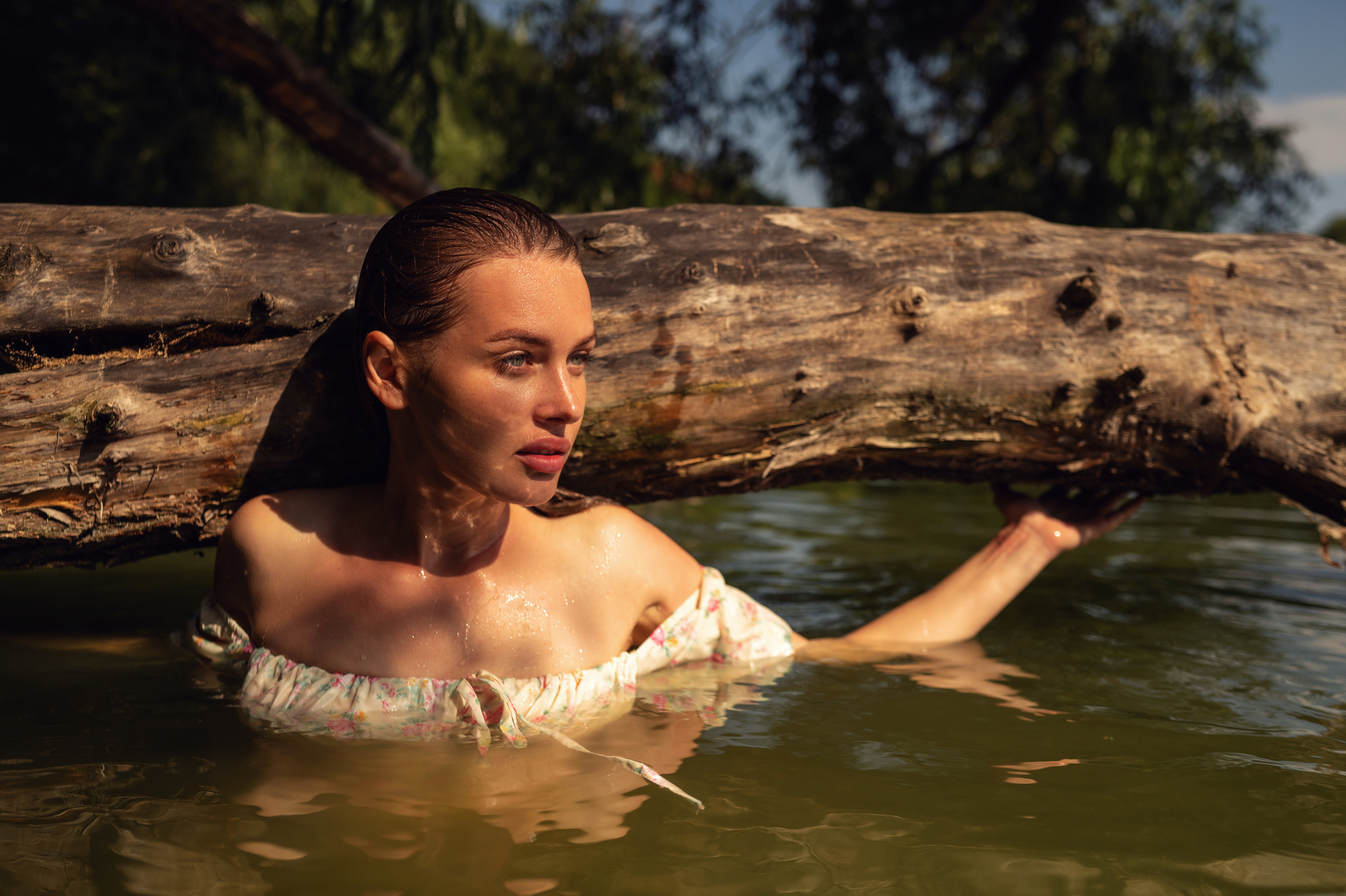 Девушка с мокрыми волосами, ярко освещена солнечным светом в воде озера возле большого ствола дерева, которое нависает над водой 