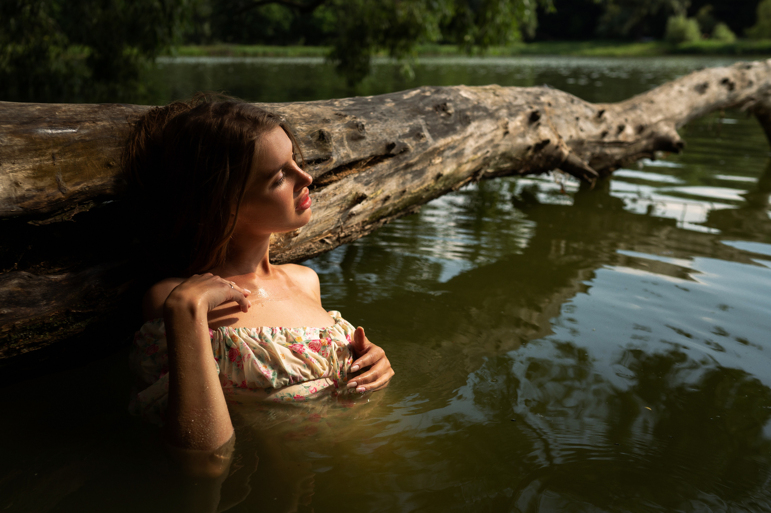 Девушка в воде озера возле большого ствола дерева, которое нависает над водой 