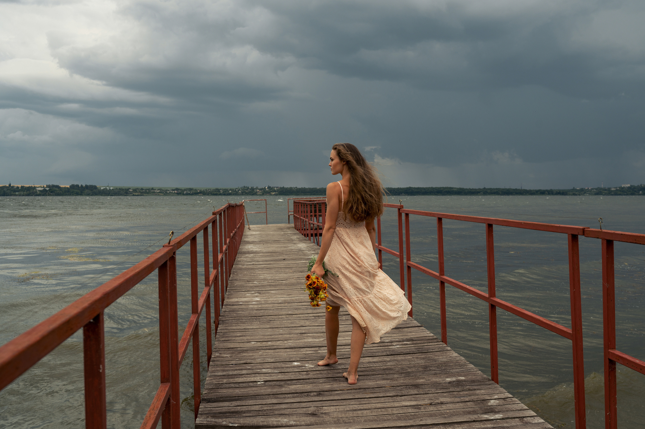 Красивая девушка в платье идет по причалу у реки на фоне грозового неба в ветренную погоду 