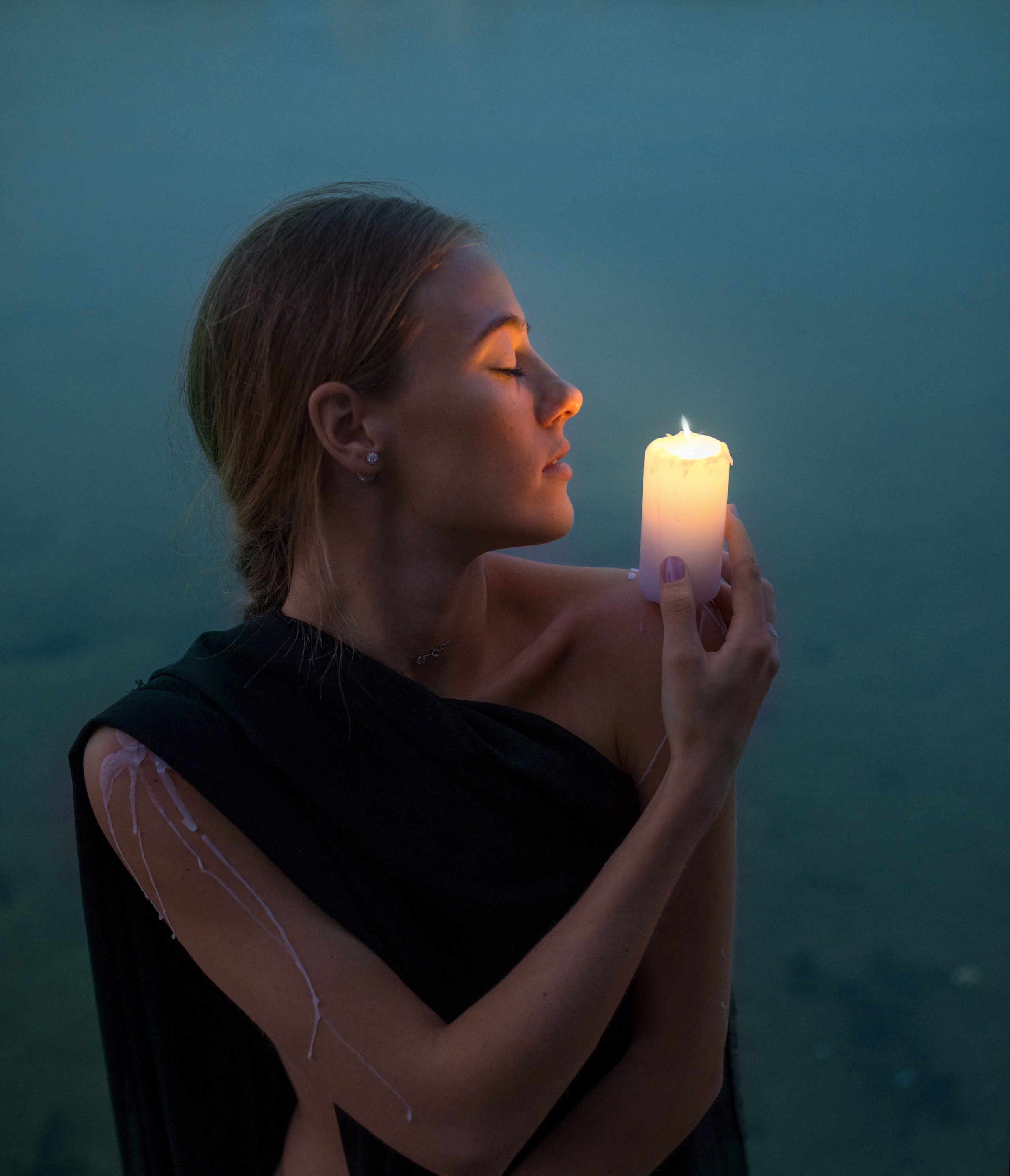 Девушка держит свечу на плече на фоне глади воды в вечернее время и смотрит в сторону горящей свечи 