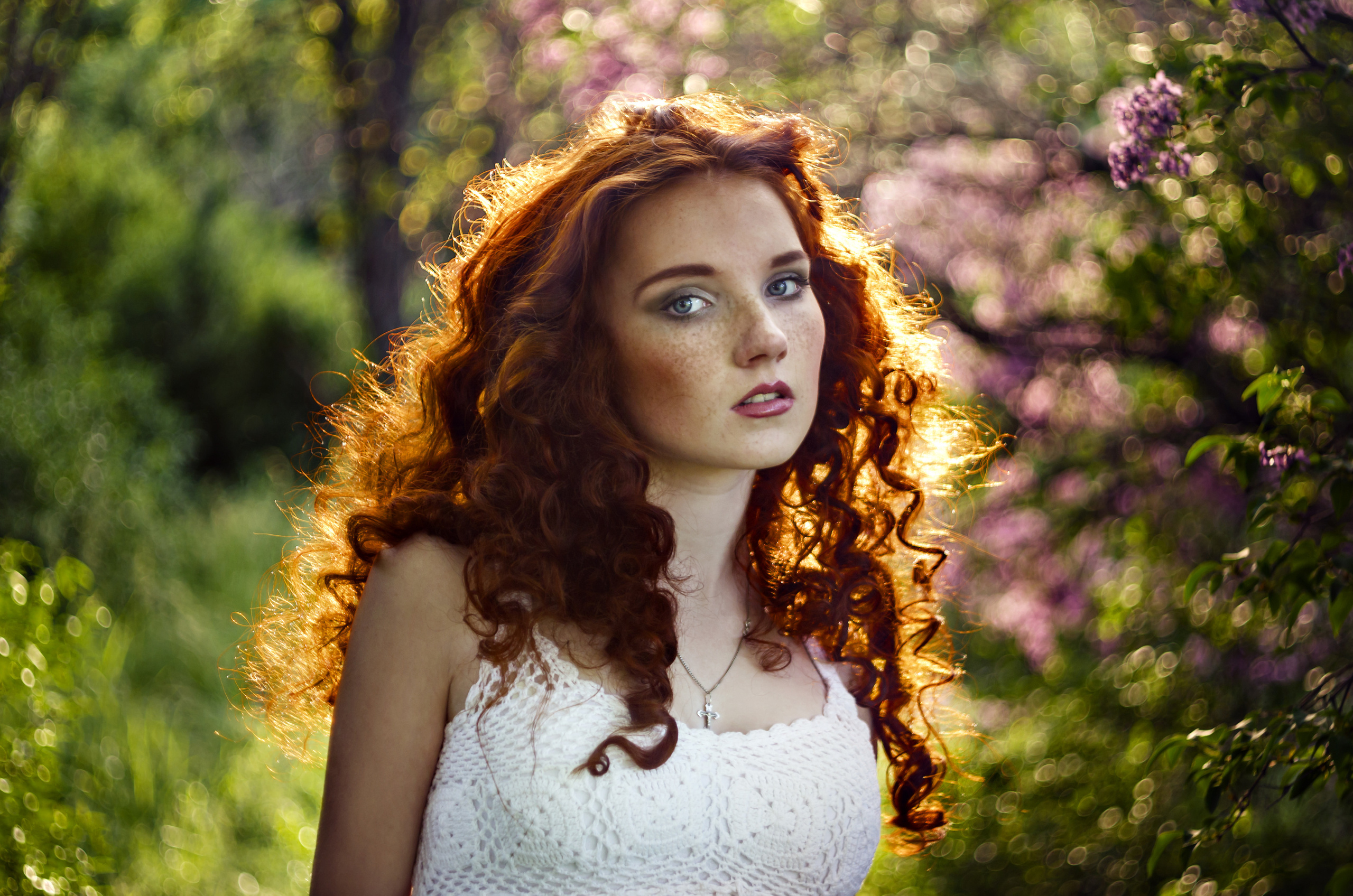 Красивая юная рыжая девушка с кудрявыми волосами в белом сарафане в контровом свете на фоне зеленой листвы и веток деревьев  
