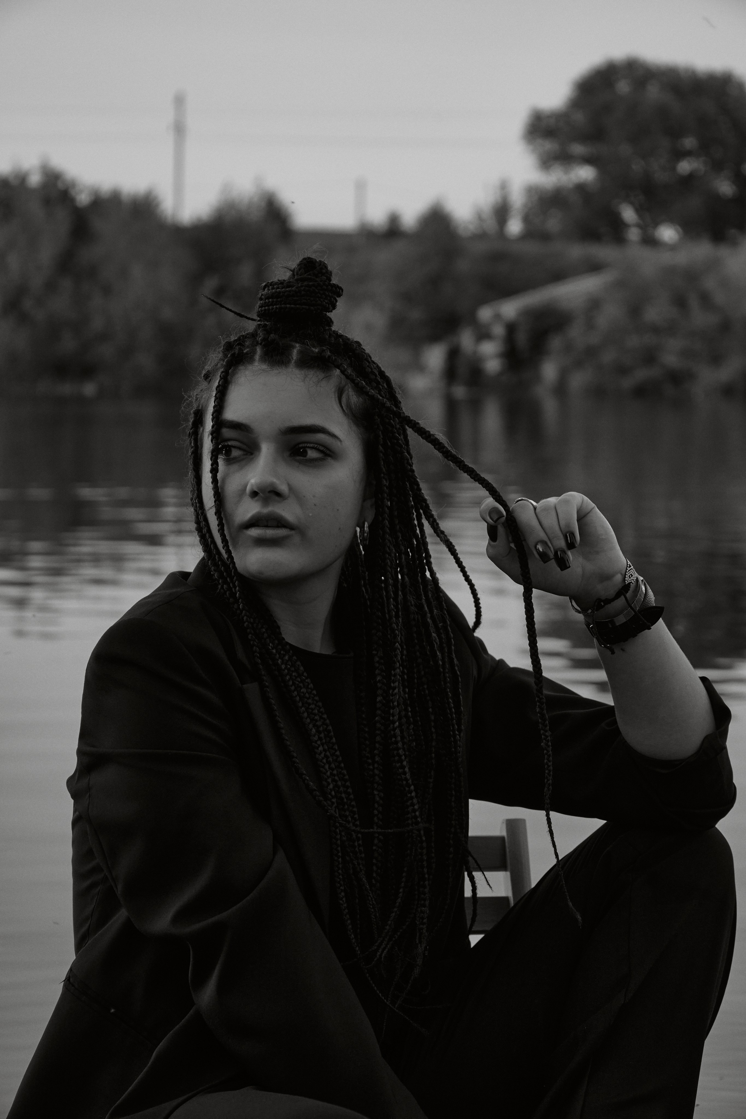 Молода жінка з афрокосами у чорному костюмі сидить на дерев'яному стільці у воді на тлі заходу сонця та зеленої природи