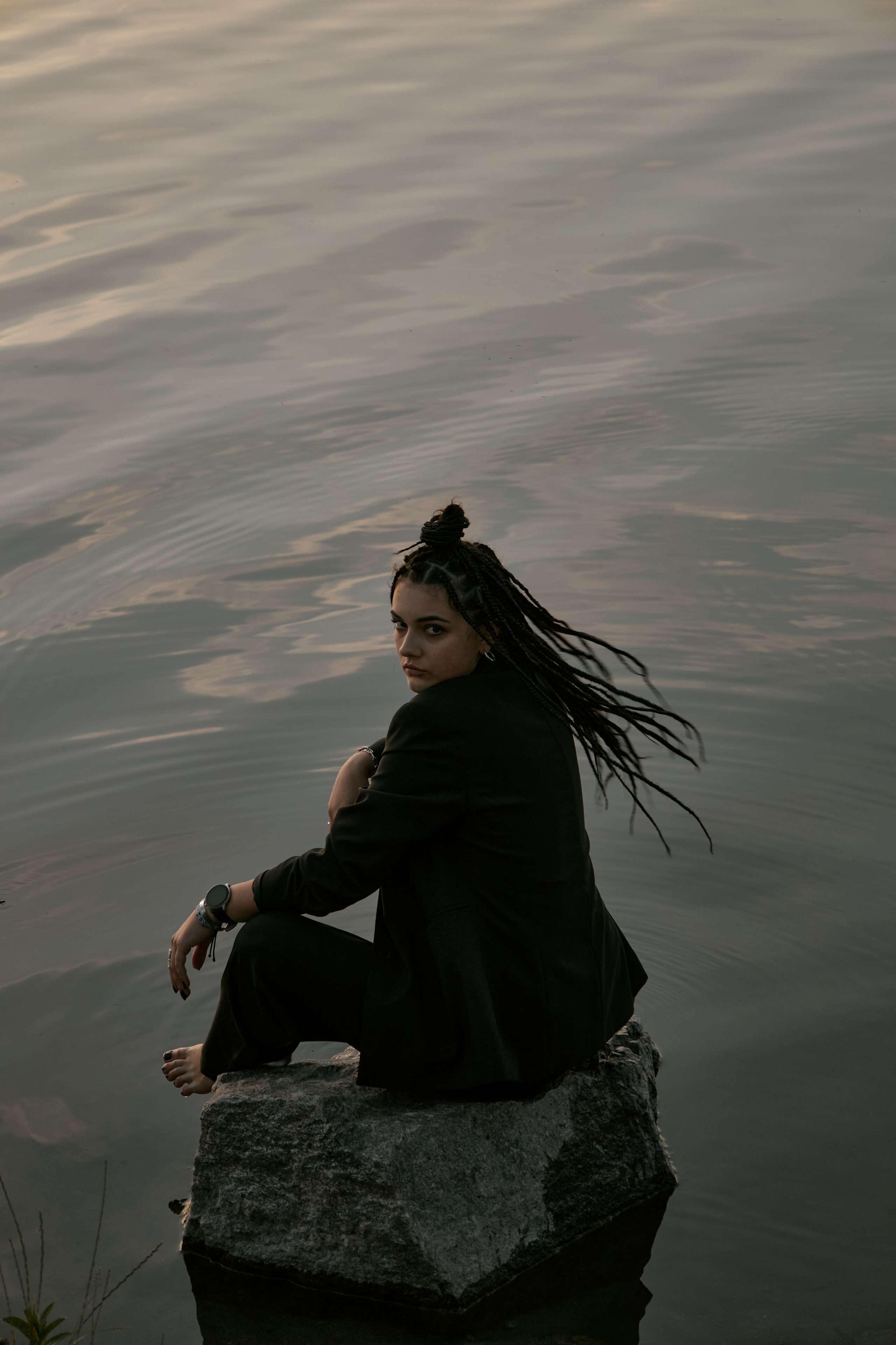 Молода жінка з афрокосами у чорному костюмі сидить на дерев'яному стільці у воді на тлі заходу сонця та зеленої природи