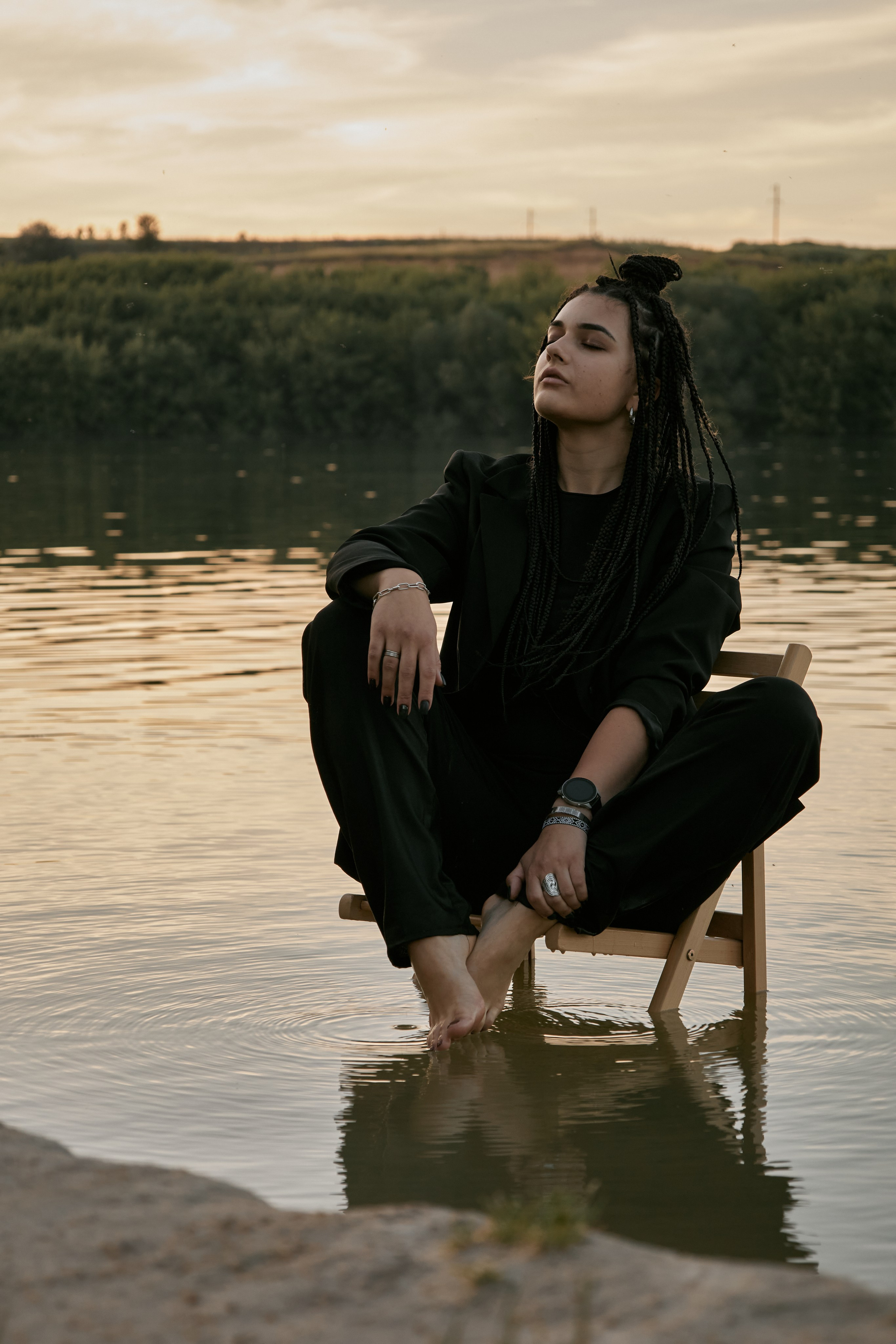 Молода жінка з афрокосами у чорному костюмі сидить на дерев'яному стільці у воді на тлі заходу сонця та зеленої природи