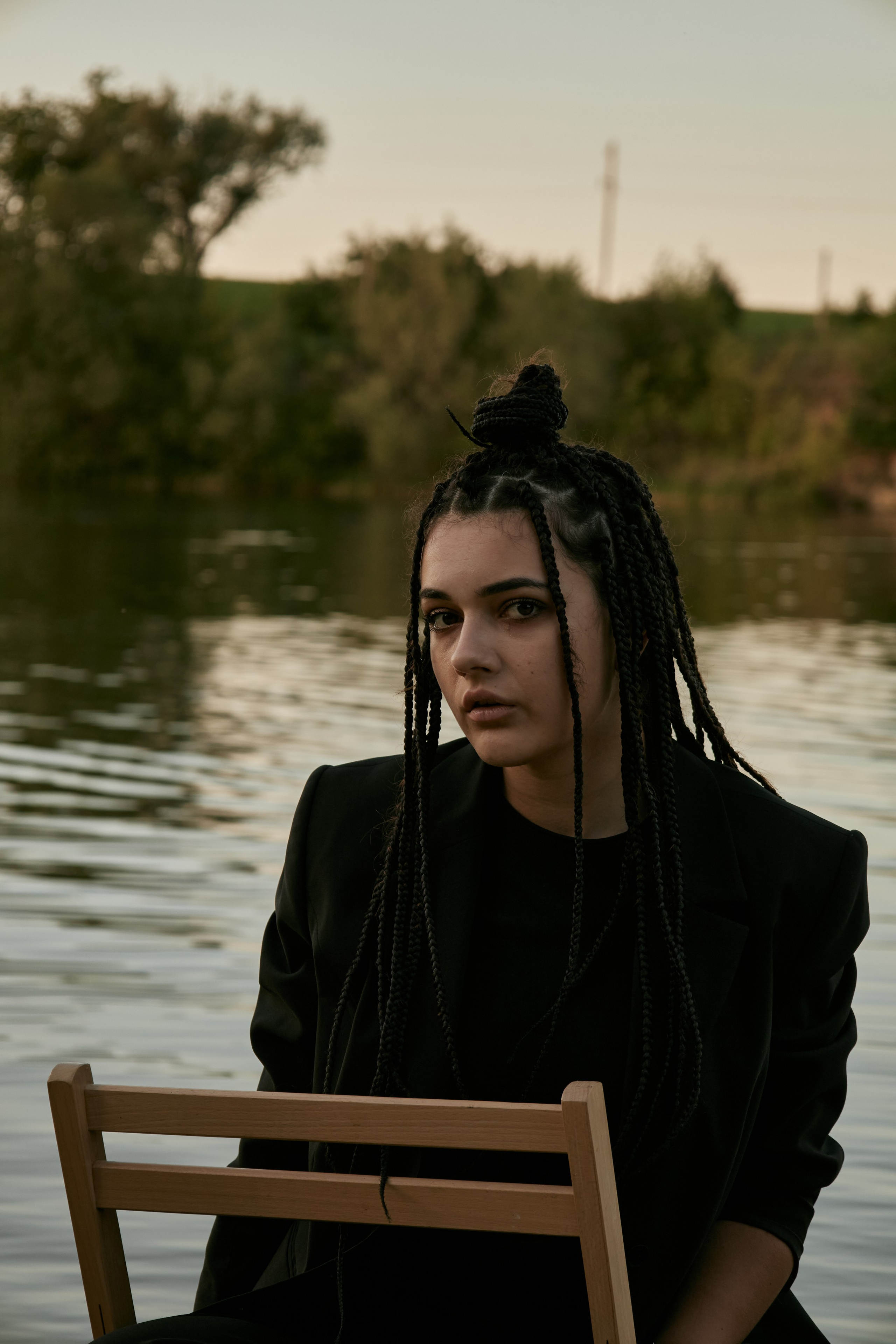 Молода жінка з афрокосами у чорному костюмі сидить на дерев'яному стільці у воді на тлі заходу сонця та зеленої природи