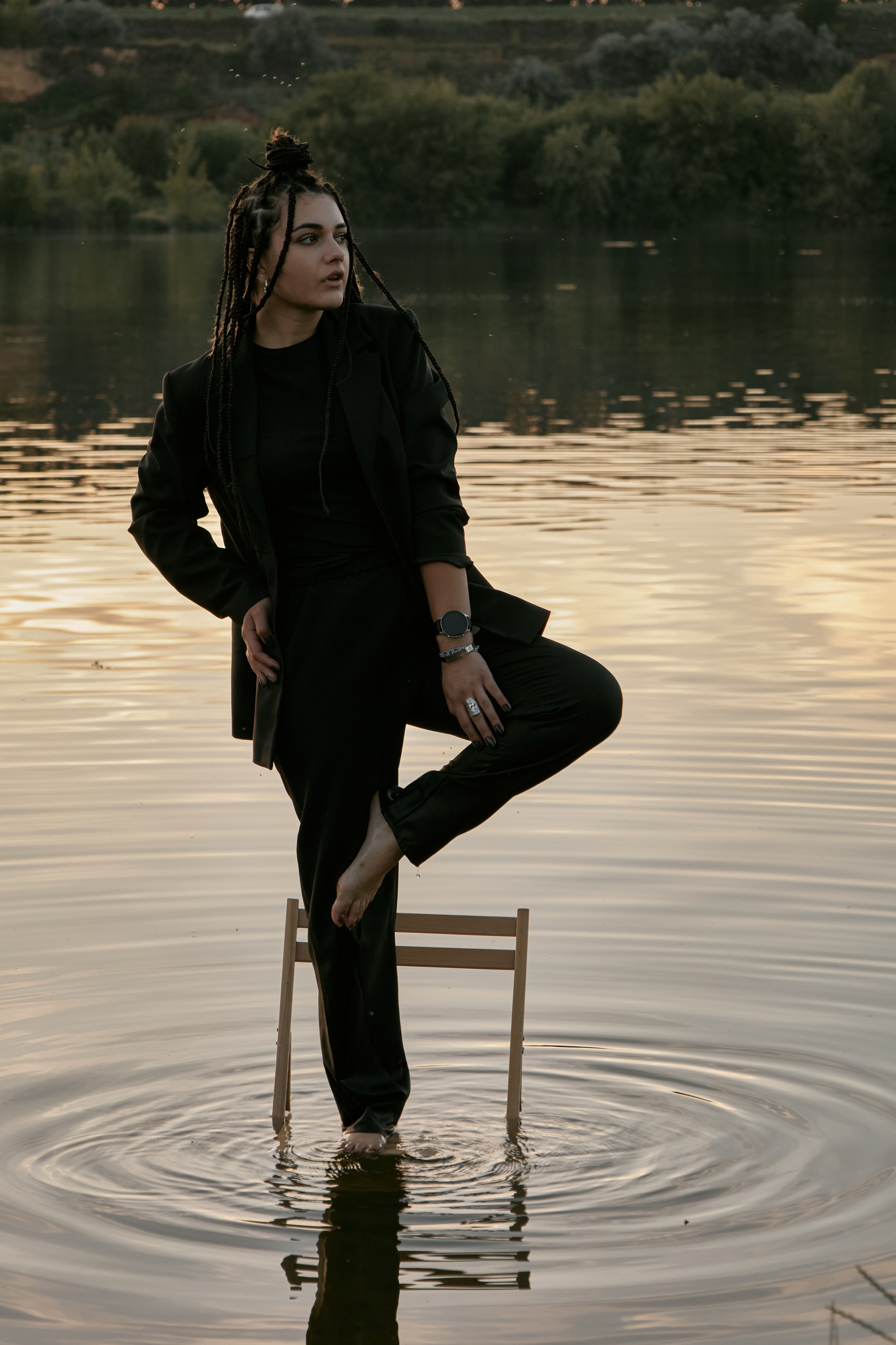 Молода жінка з афрокосами у чорному костюмі сидить на дерев'яному стільці у воді на тлі заходу сонця та зеленої природи