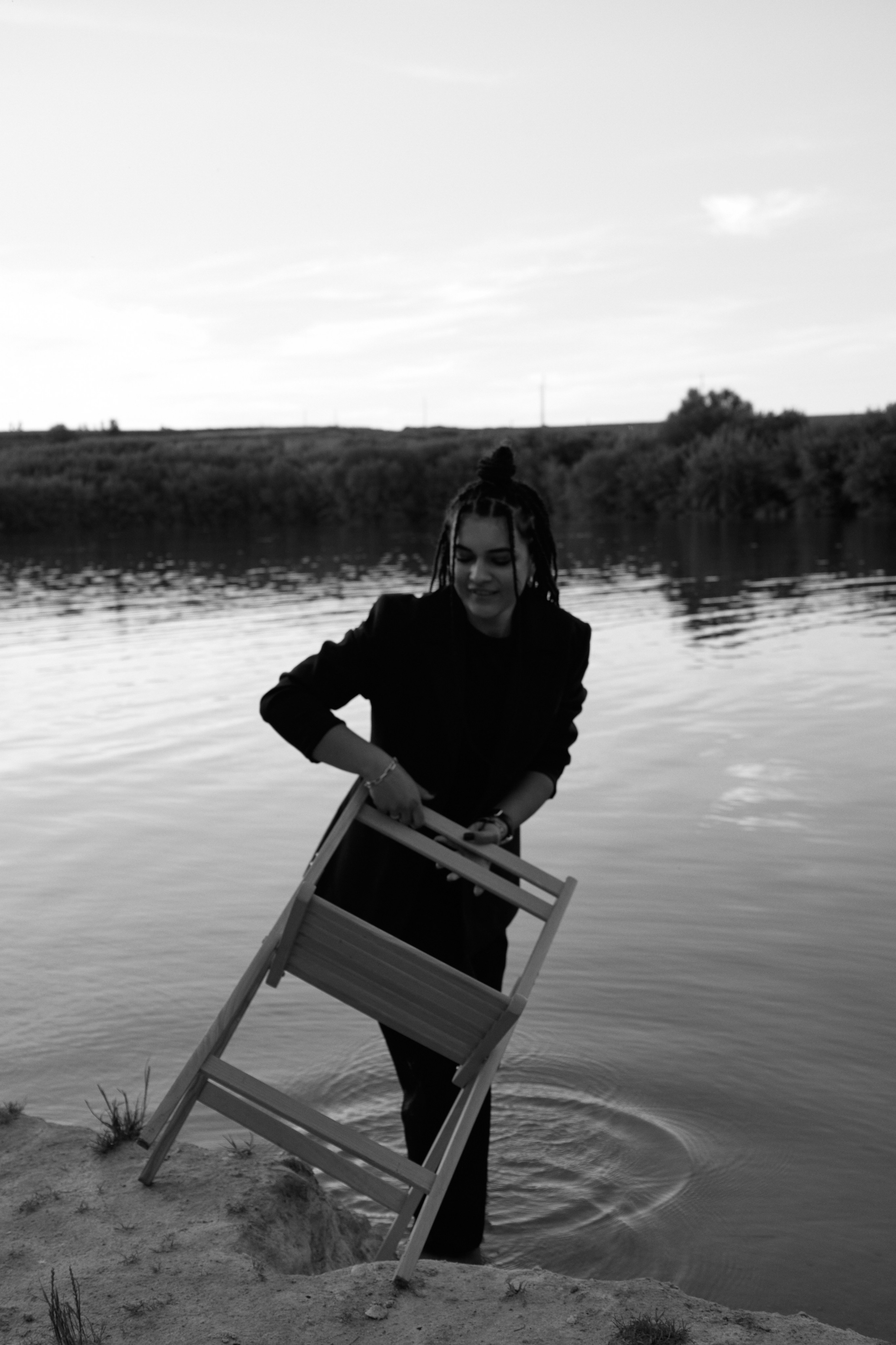 Молода жінка з афрокосами у чорному костюмі сидить на дерев'яному стільці у воді на тлі заходу сонця та зеленої природи