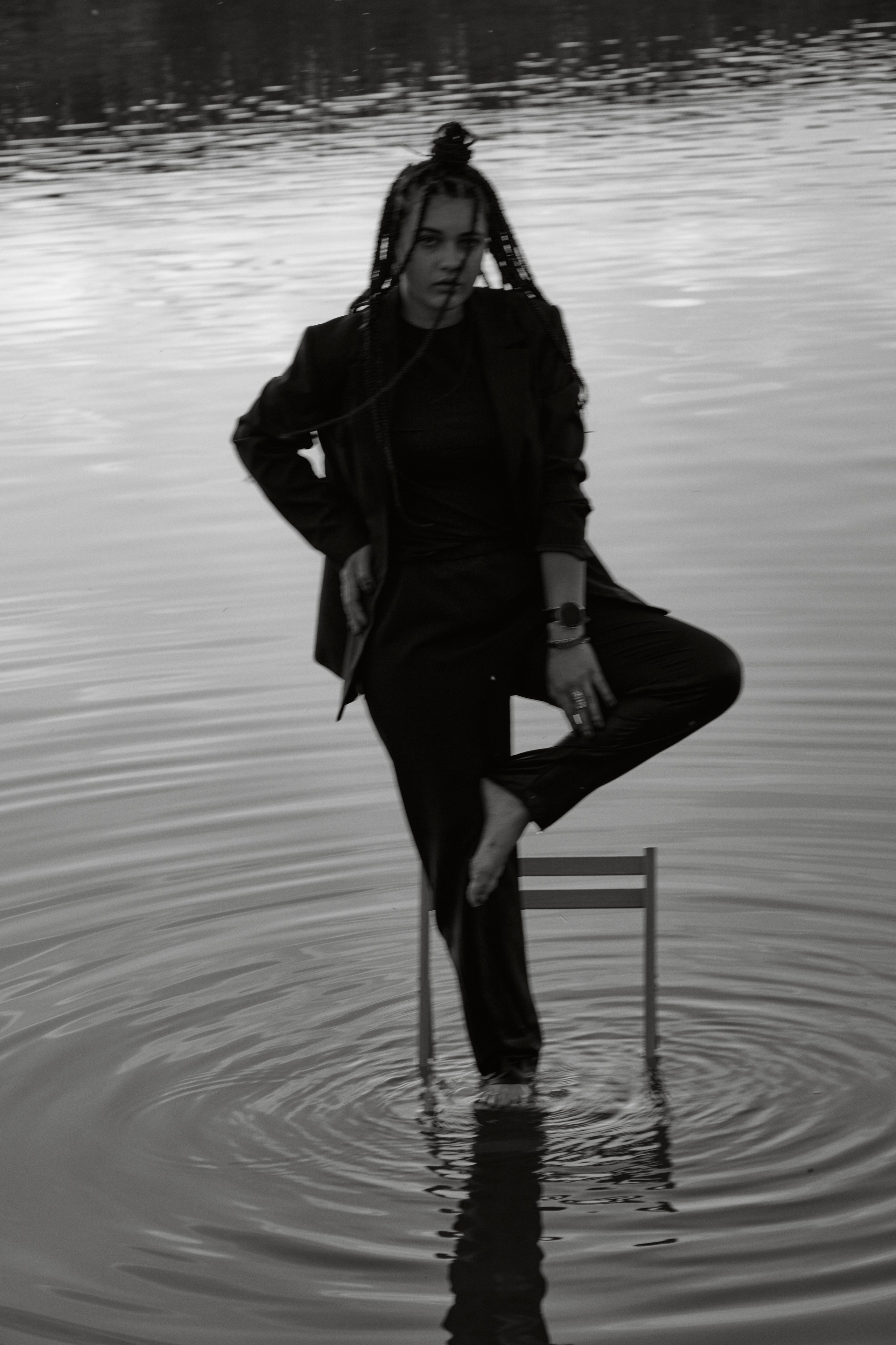 Молода жінка з афрокосами у чорному костюмі сидить на дерев'яному стільці у воді на тлі заходу сонця та зеленої природи