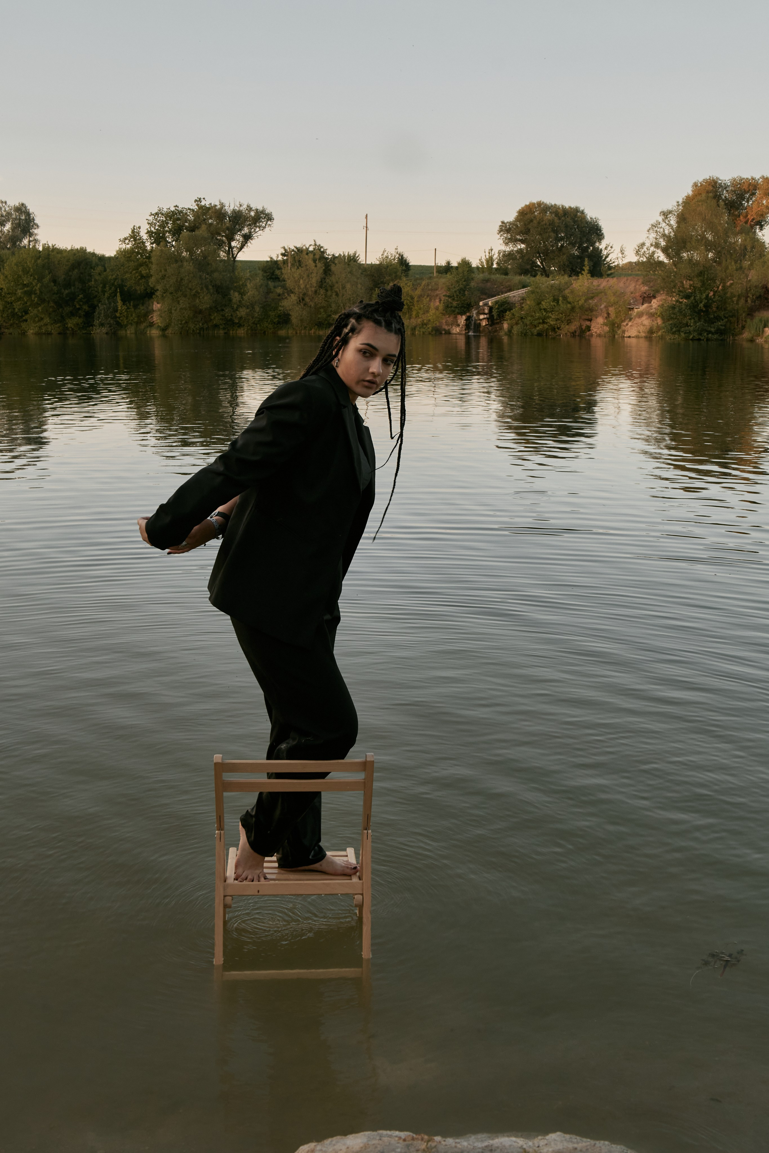 Молода жінка з афрокосами у чорному костюмі сидить на дерев'яному стільці у воді на тлі заходу сонця та зеленої природи