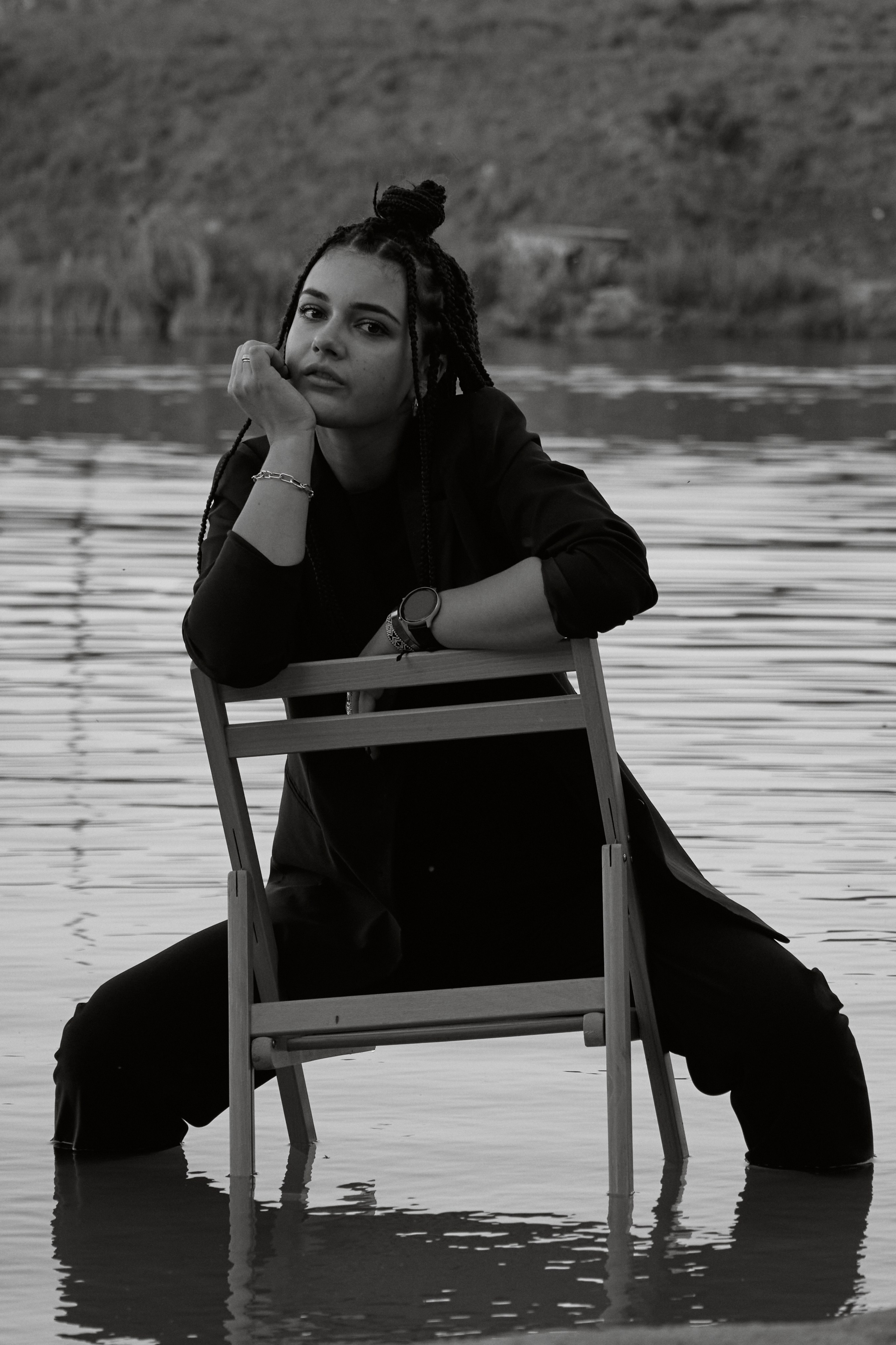 Молода жінка з афрокосами у чорному костюмі сидить на дерев'яному стільці у воді на тлі заходу сонця та зеленої природи