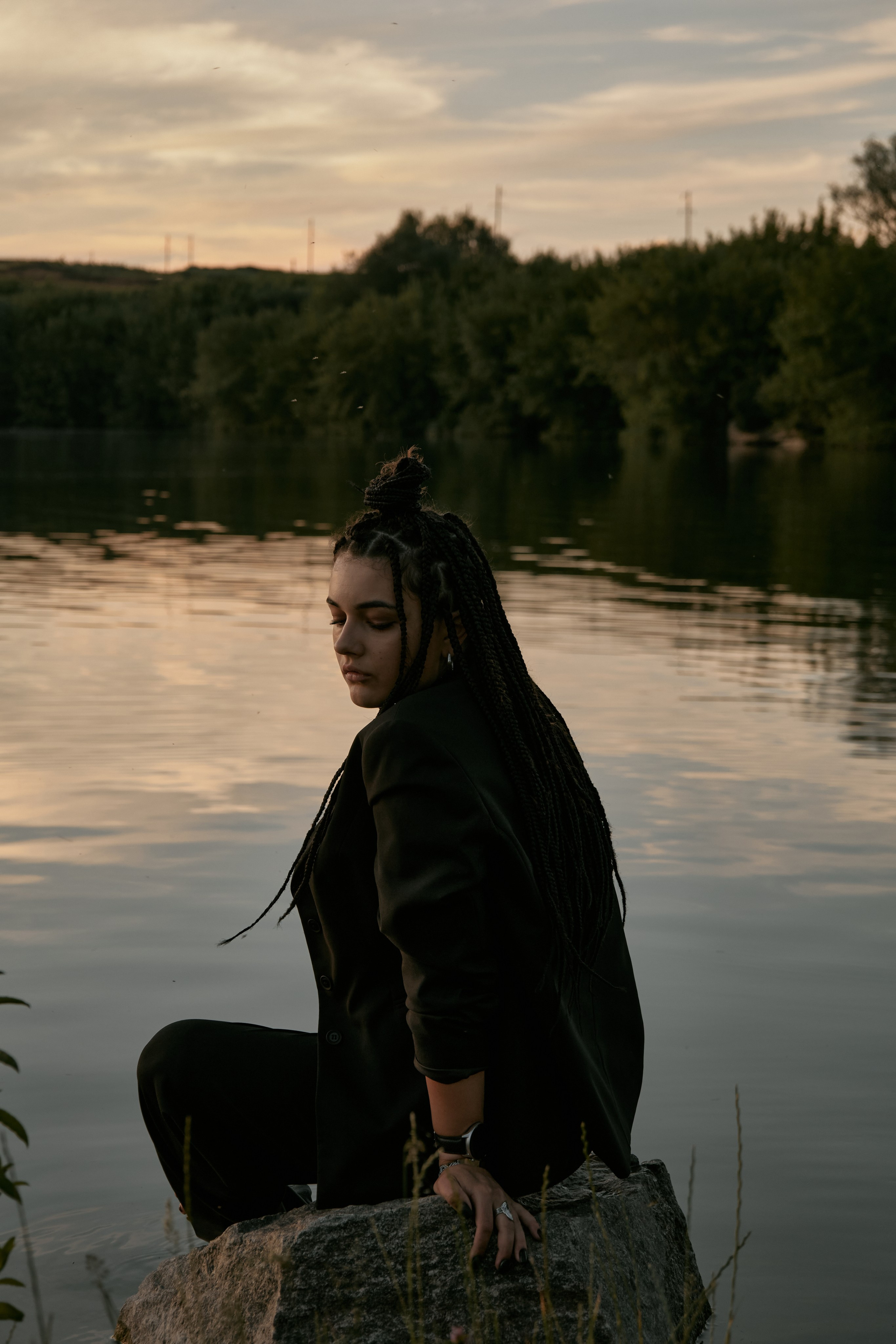 Молода жінка з афрокосами у чорному костюмі сидить на дерев'яному стільці у воді на тлі заходу сонця та зеленої природи