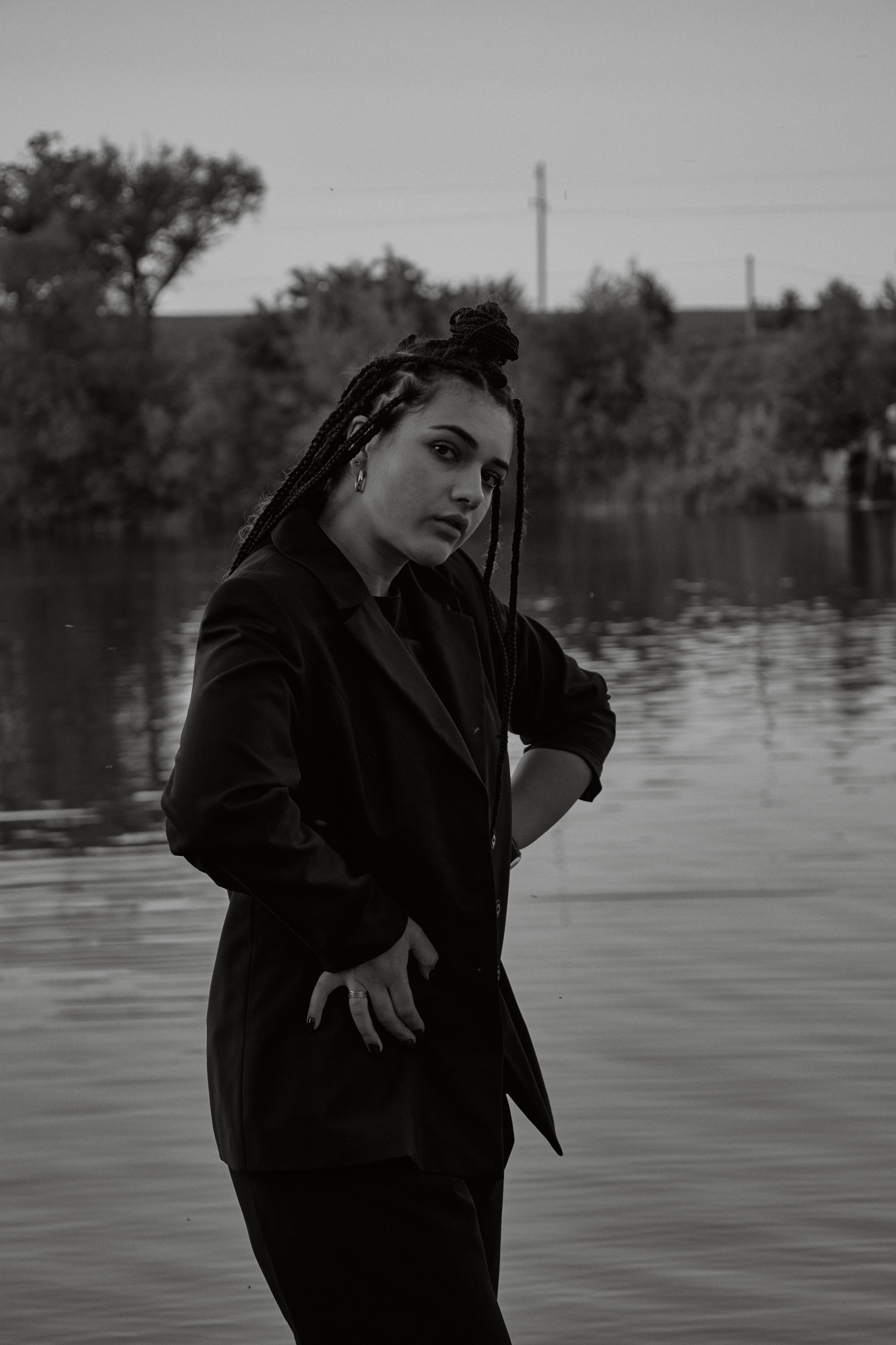 Молода жінка з афрокосами у чорному костюмі сидить на дерев'яному стільці у воді на тлі заходу сонця та зеленої природи