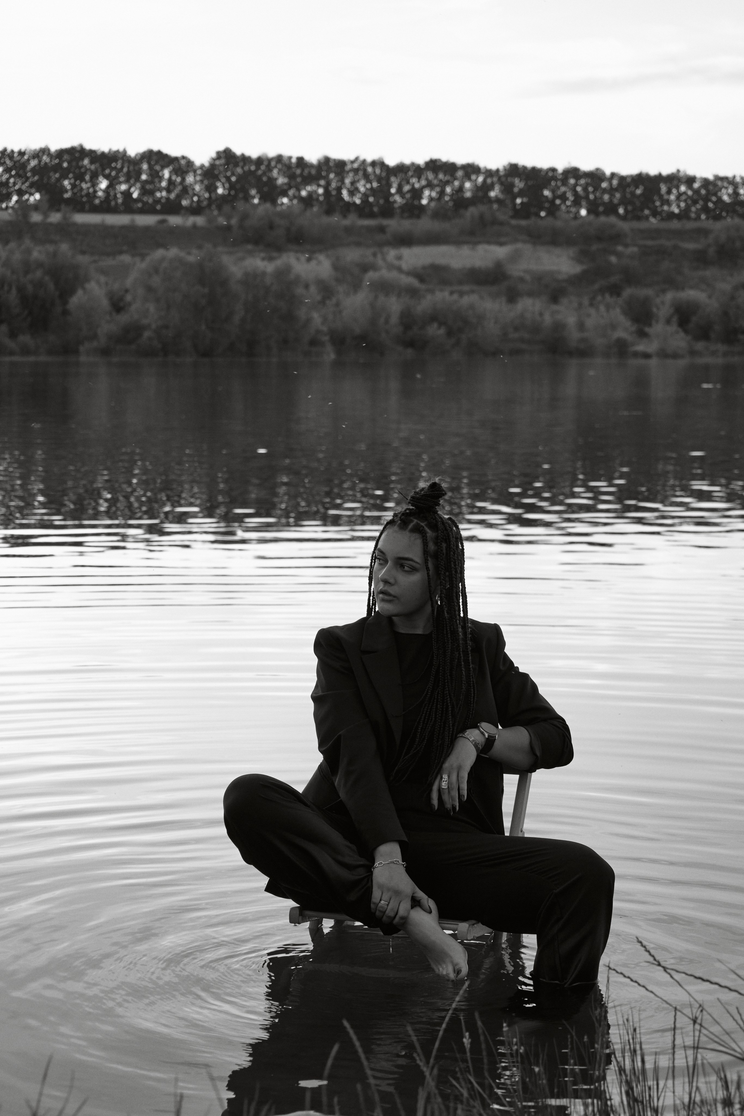 Молода жінка з афрокосами у чорному костюмі сидить на дерев'яному стільці у воді на тлі заходу сонця та зеленої природи