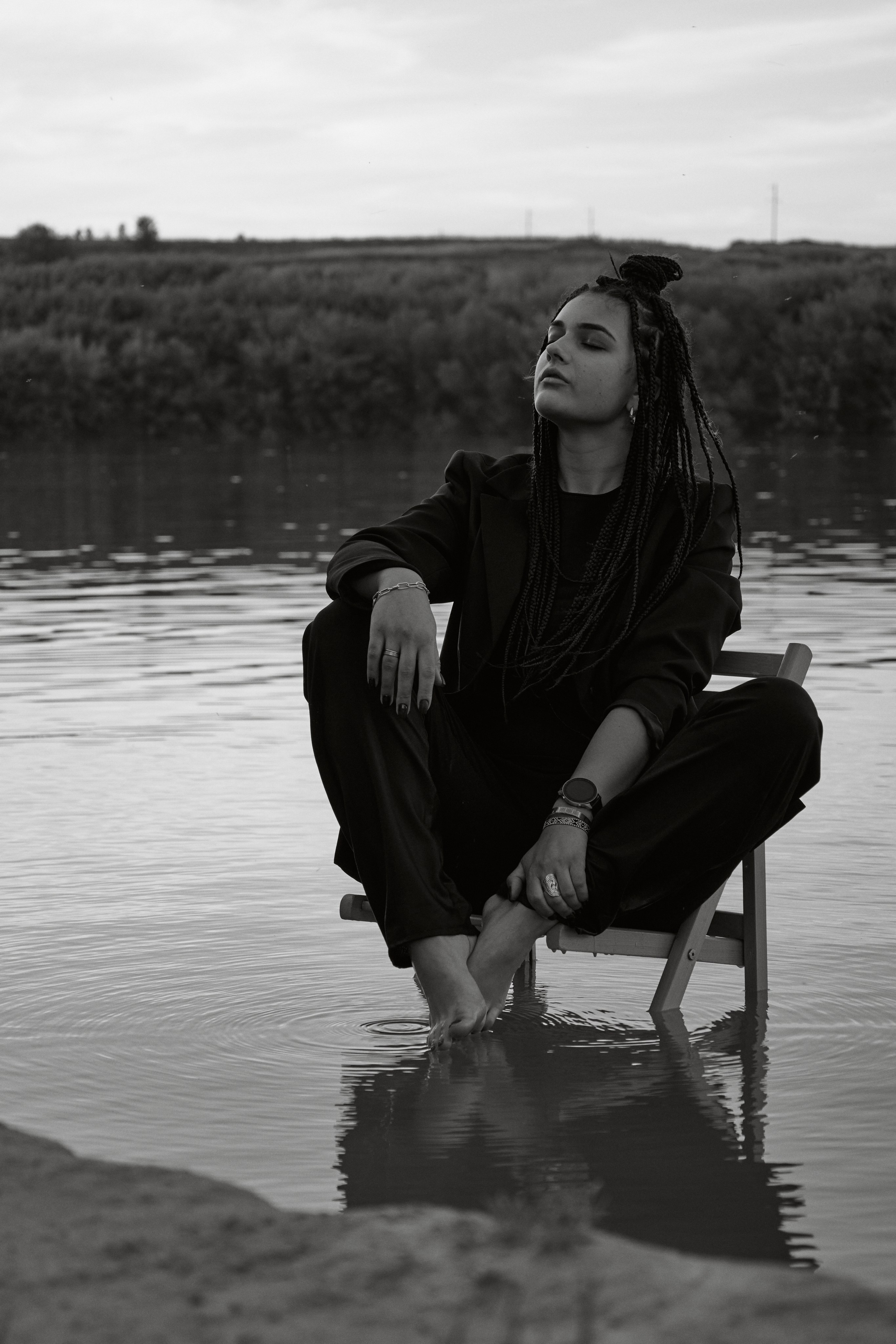 Молода жінка з афрокосами у чорному костюмі сидить на дерев'яному стільці у воді на тлі заходу сонця та зеленої природи