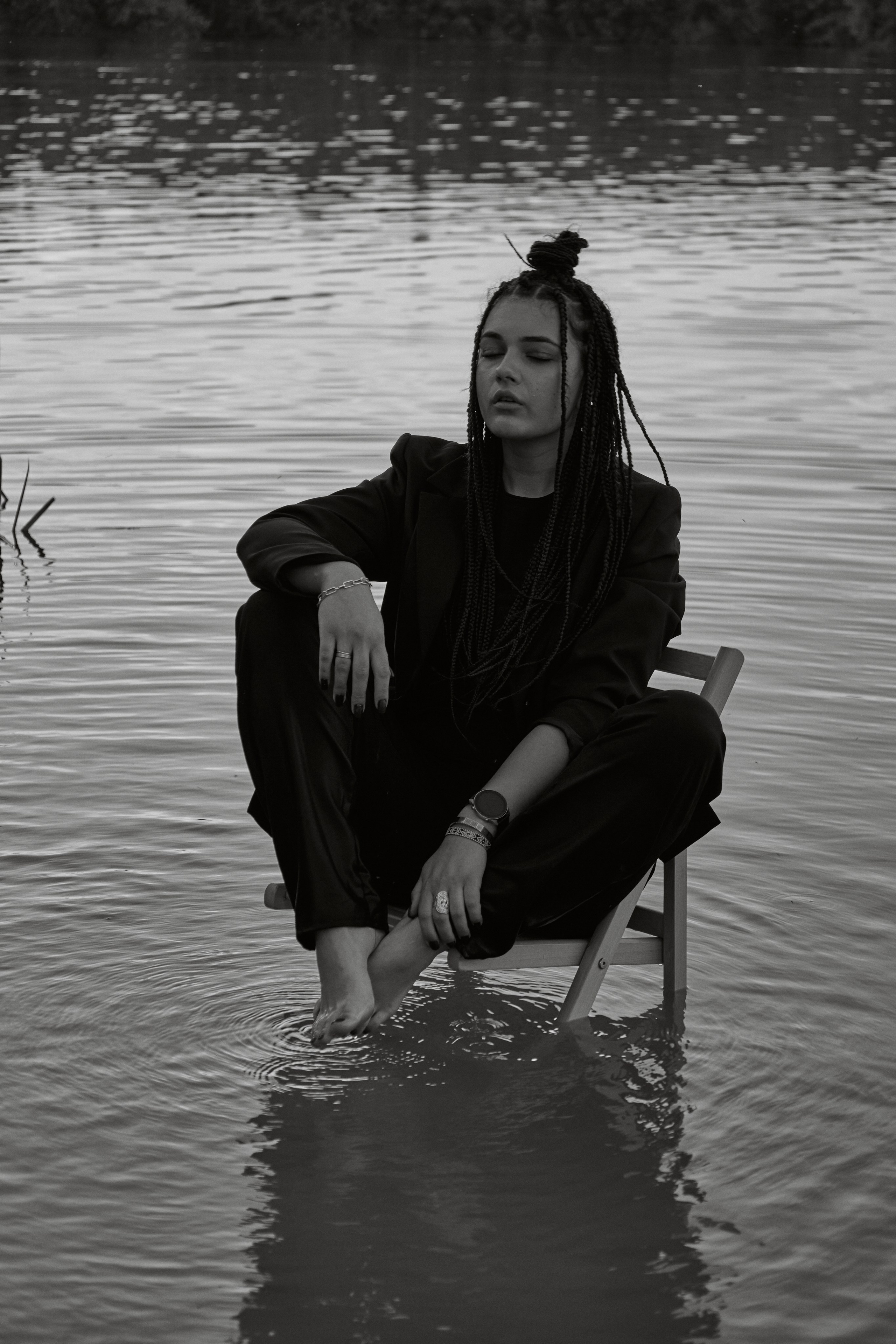 Молода жінка з афрокосами у чорному костюмі сидить на дерев'яному стільці у воді на тлі заходу сонця та зеленої природи