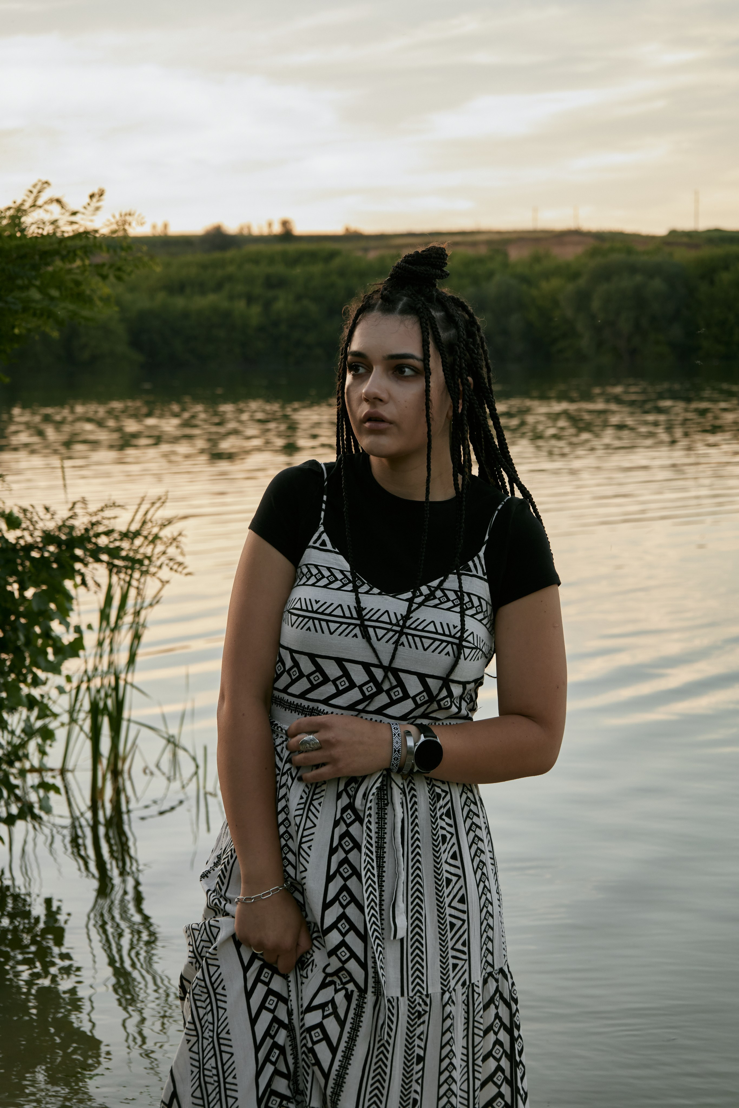 Молода жінка з афрокосами у чорному костюмі сидить на дерев'яному стільці у воді на тлі заходу сонця та зеленої природи