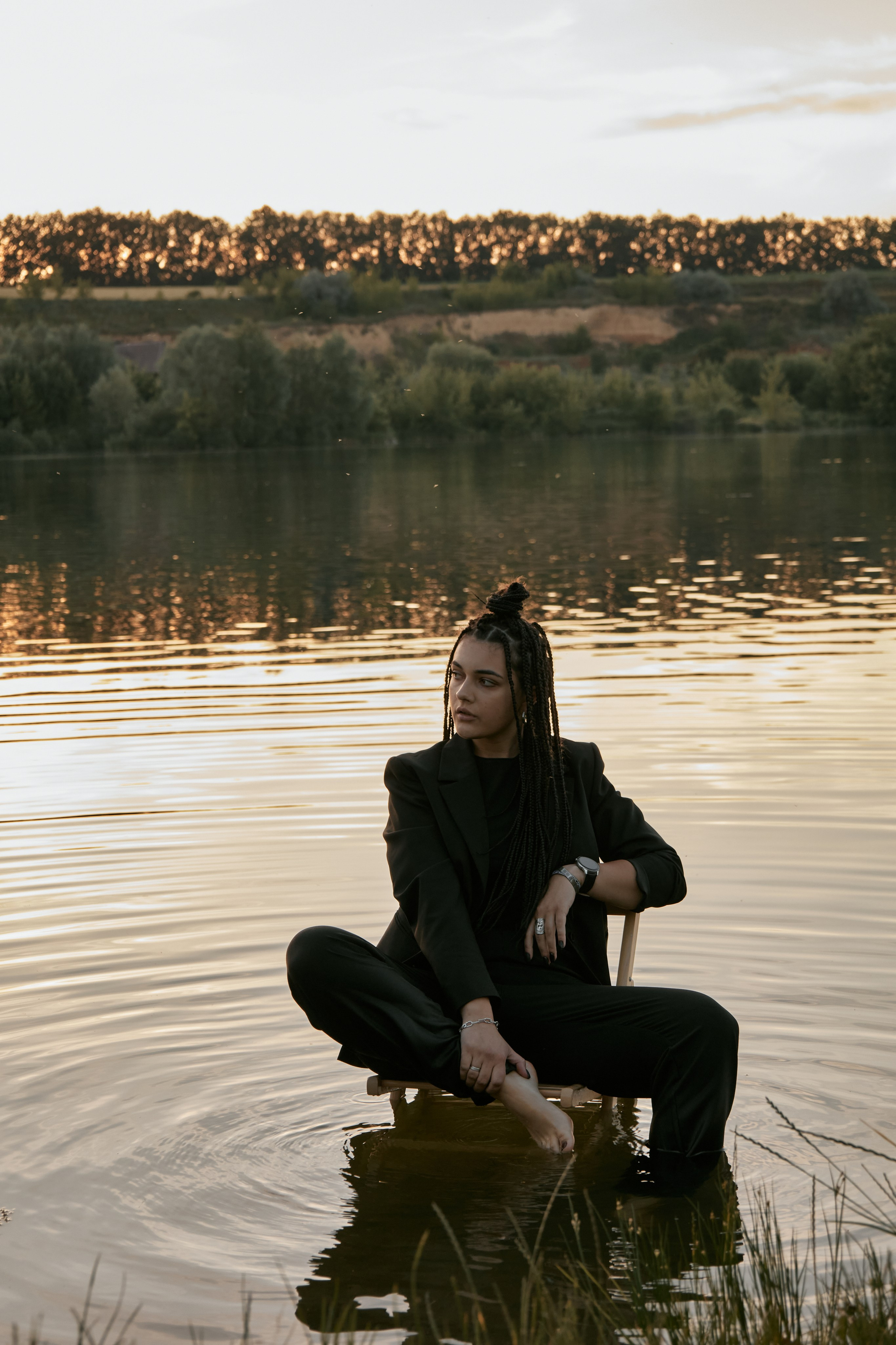 Молода жінка з афрокосами у чорному костюмі сидить на дерев'яному стільці у воді на тлі заходу сонця та зеленої природи