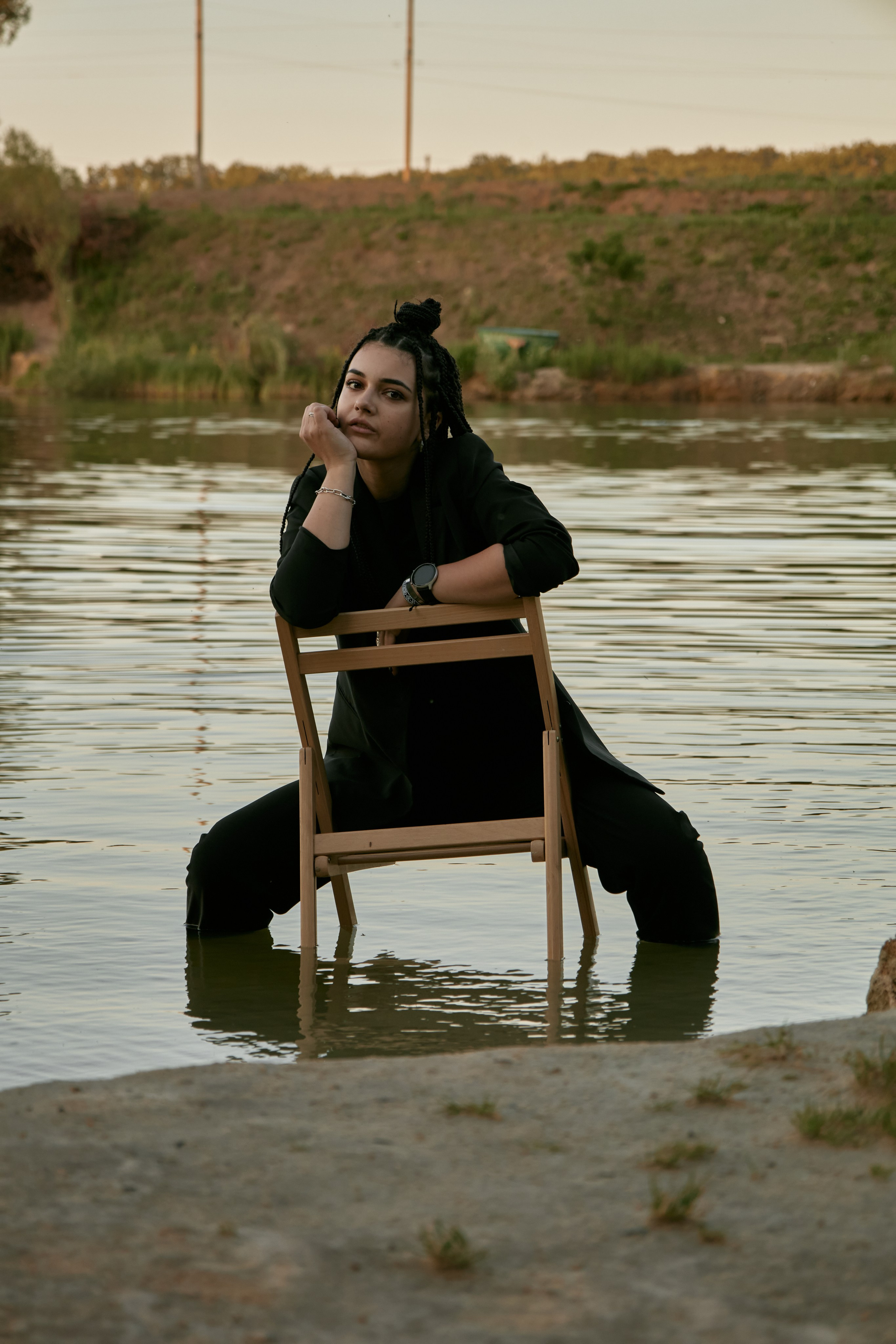 Молода жінка з афрокосами у чорному костюмі сидить на дерев'яному стільці у воді на тлі заходу сонця та зеленої природи