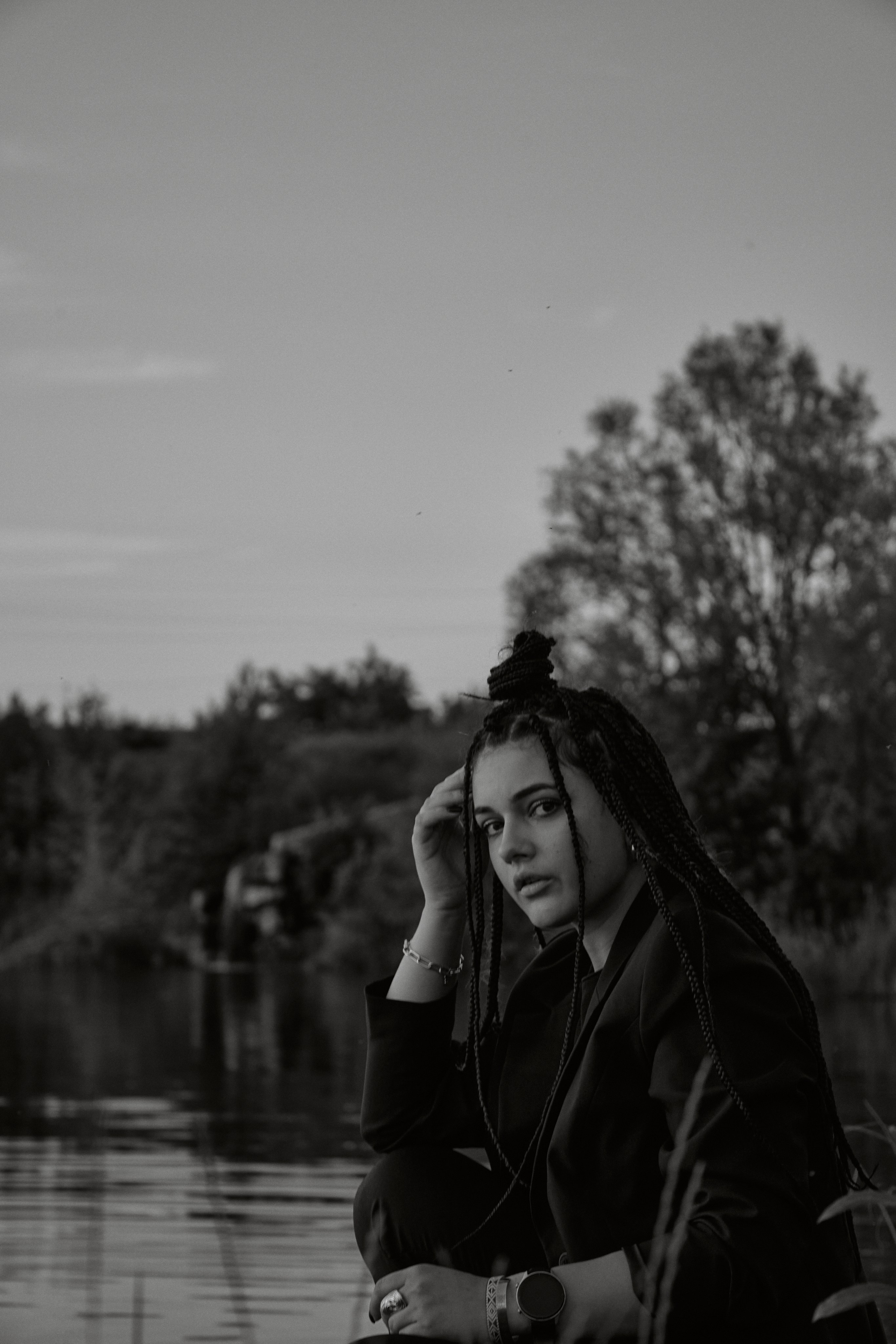 Молода жінка з афрокосами у чорному костюмі сидить на дерев'яному стільці у воді на тлі заходу сонця та зеленої природи