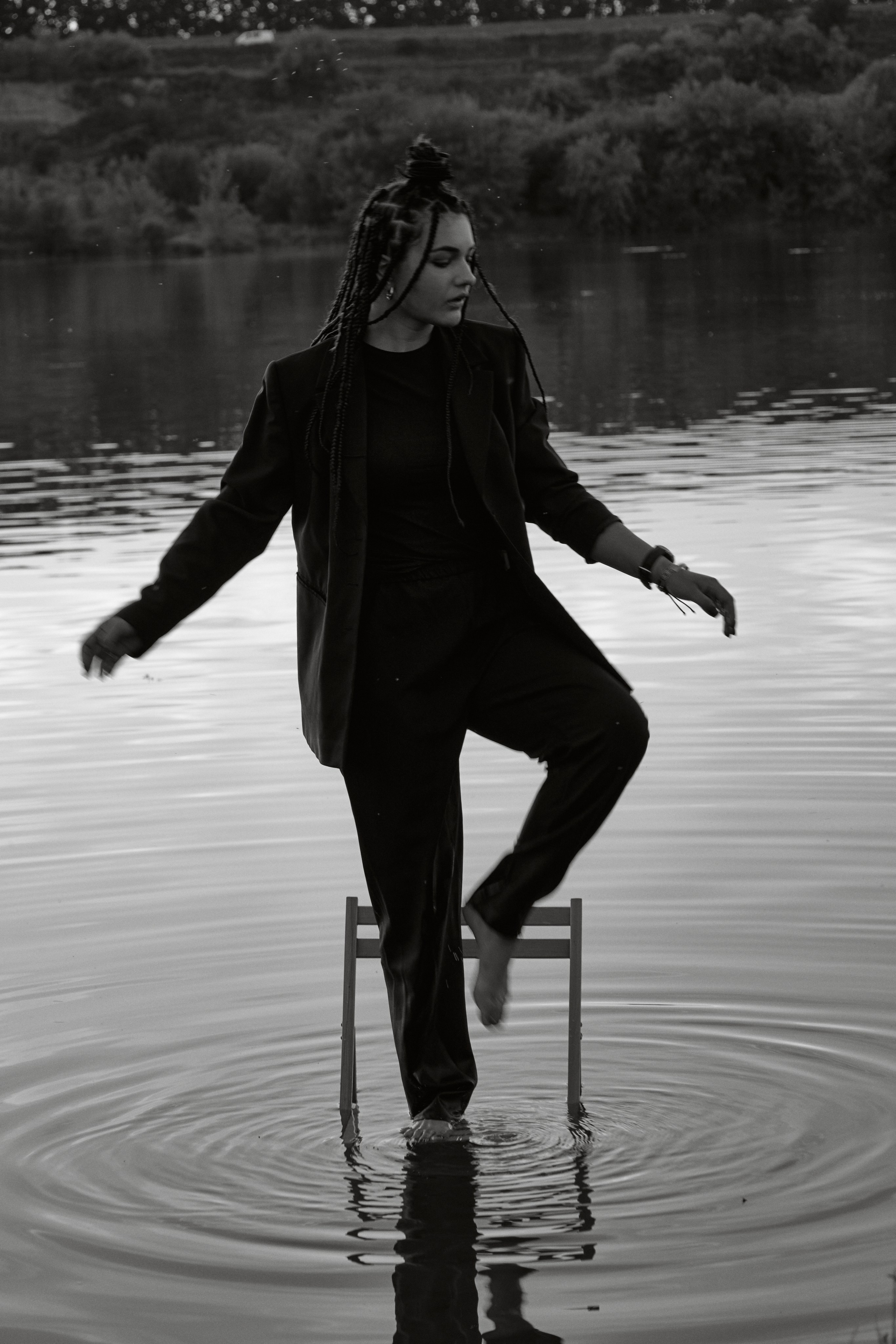 Молода жінка з афрокосами у чорному костюмі сидить на дерев'яному стільці у воді на тлі заходу сонця та зеленої природи