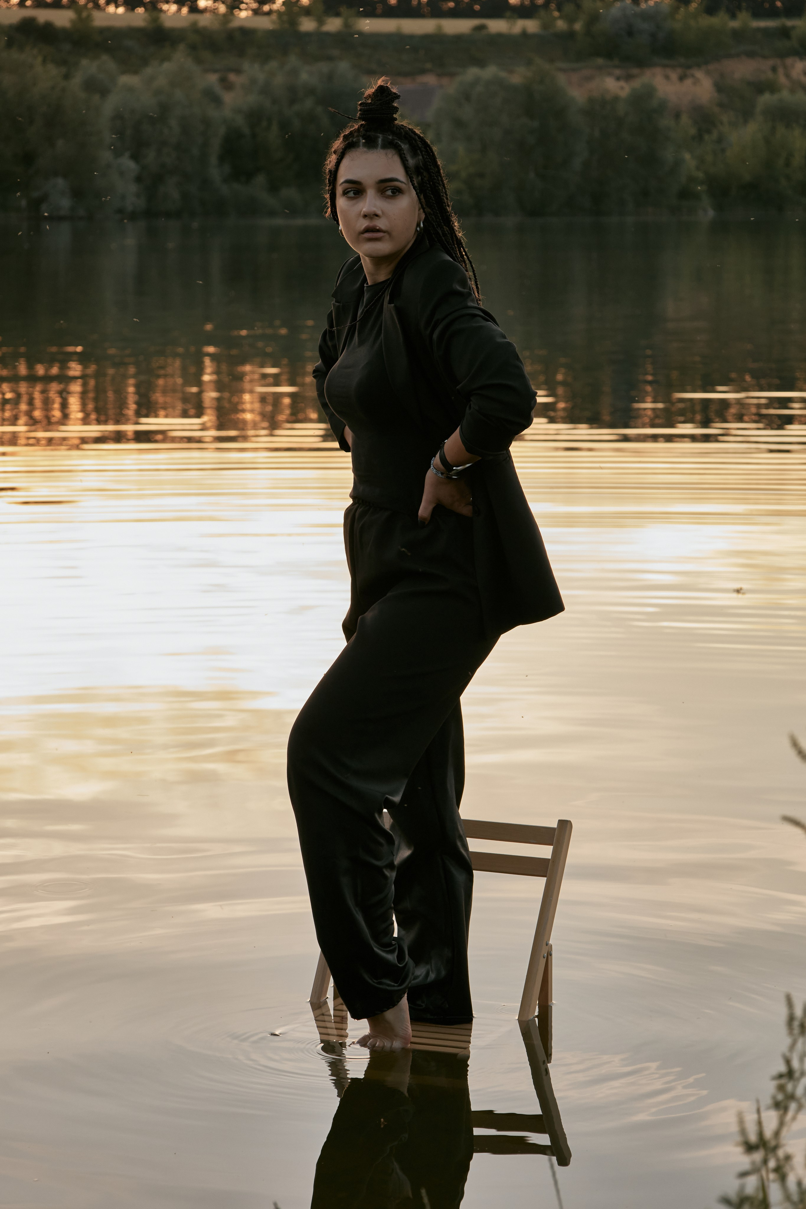 Молода жінка з афрокосами у чорному костюмі сидить на дерев'яному стільці у воді на тлі заходу сонця та зеленої природи