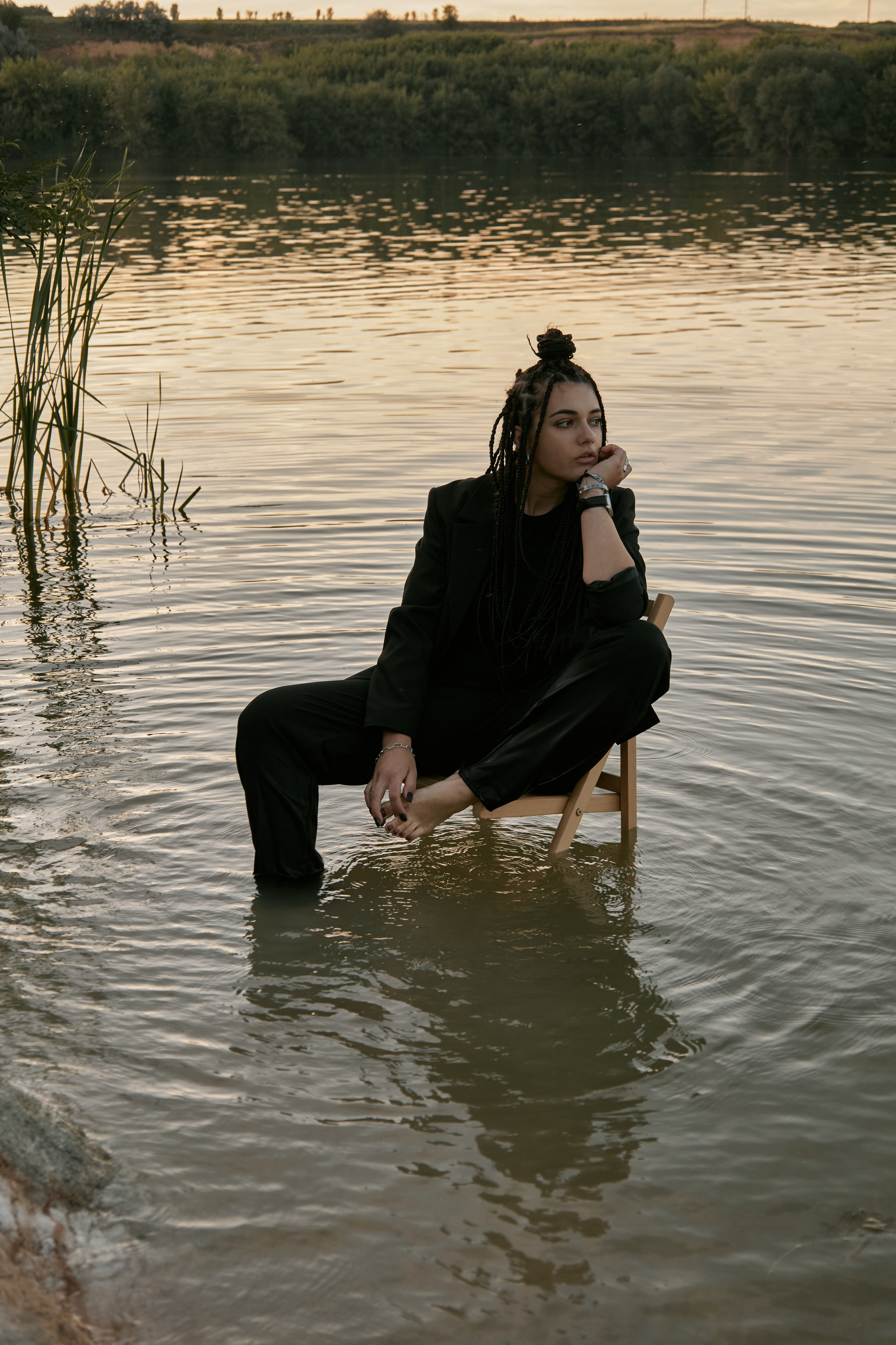Молода жінка з афрокосами у чорному костюмі сидить на дерев'яному стільці у воді на тлі заходу сонця та зеленої природи