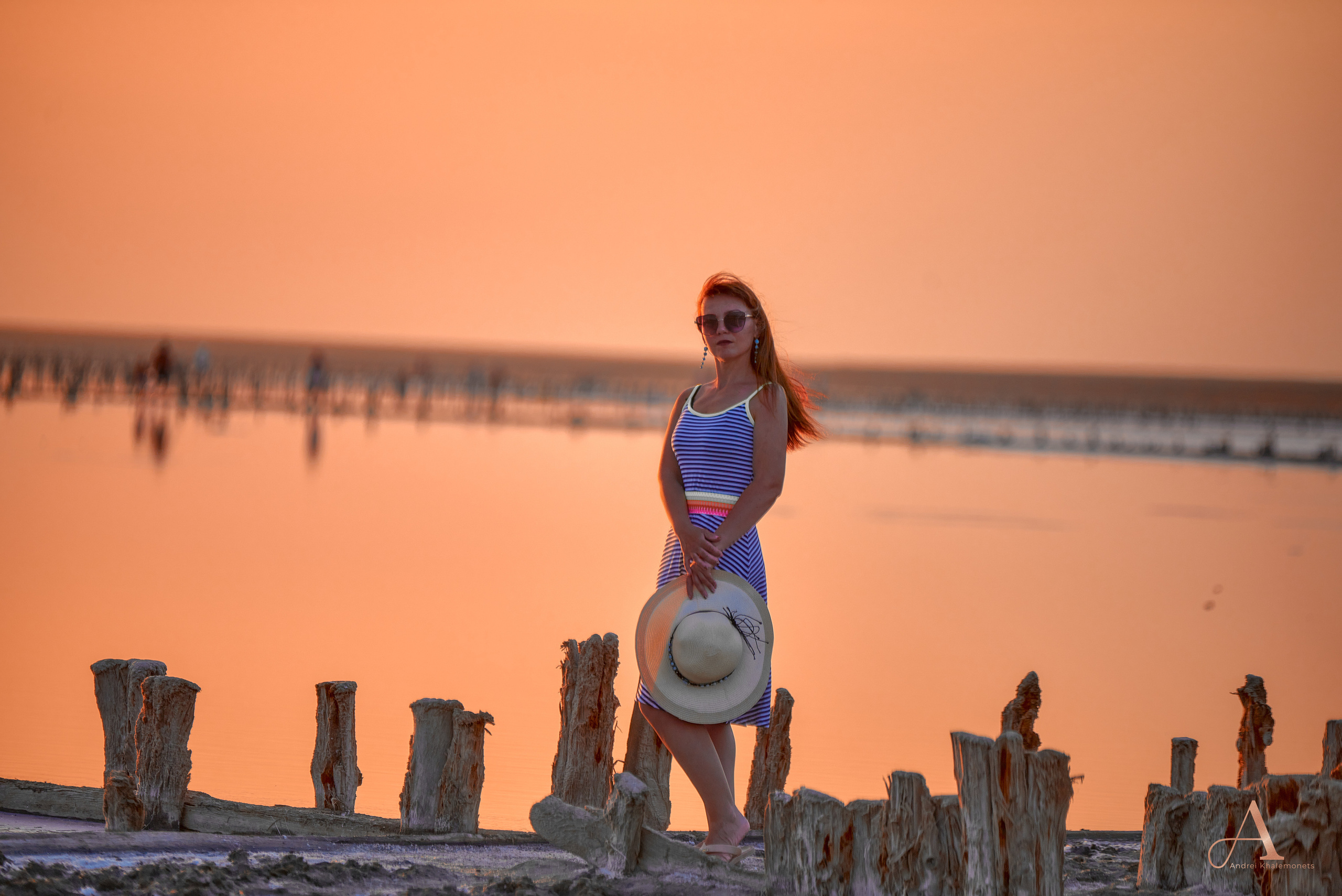 Девушка в платье с большой соломенной шляпой в руках на закате у озера.