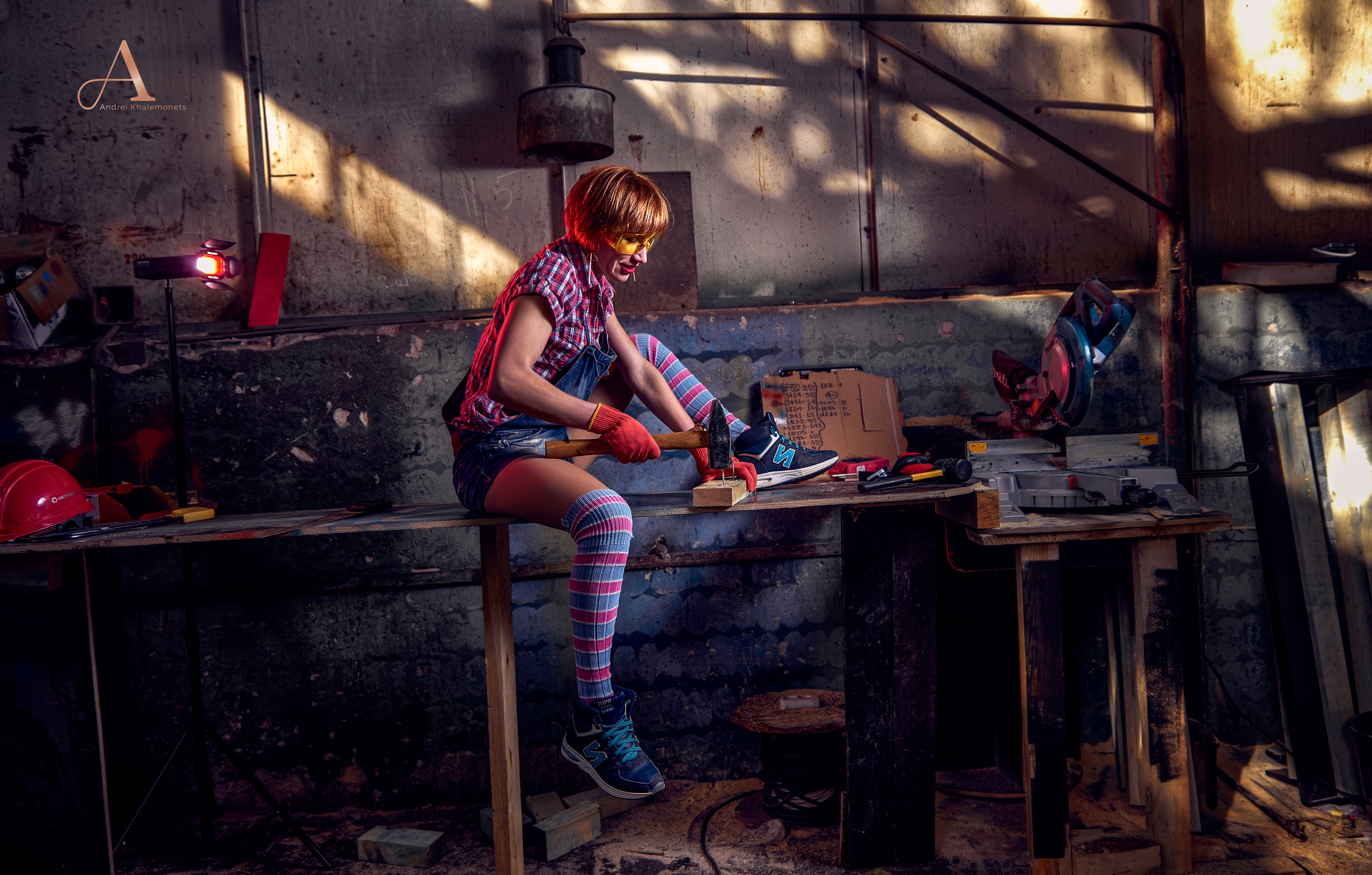 Девушка в джинсовом комбинезоне с молотком в руках сидит на стеллаже.