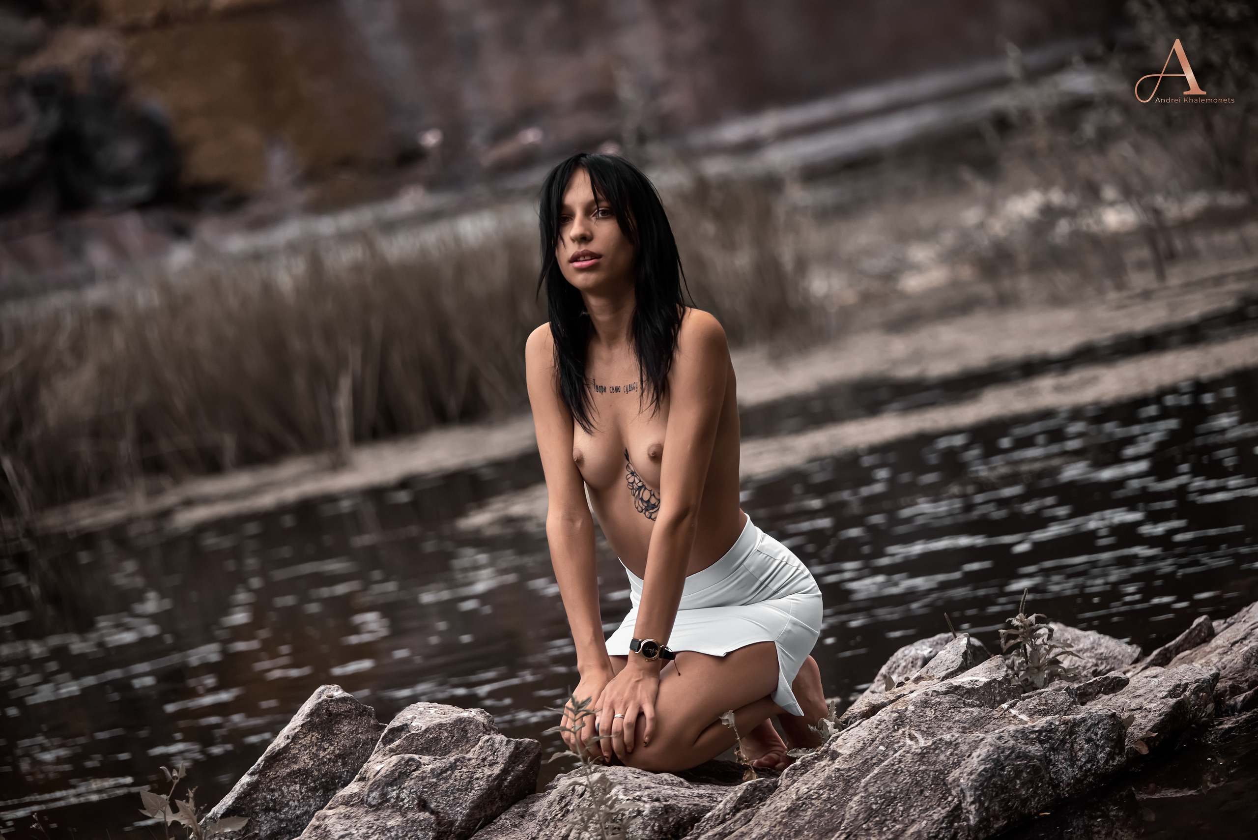 Красивая юная девушка позирует топлес на камнях у воды.