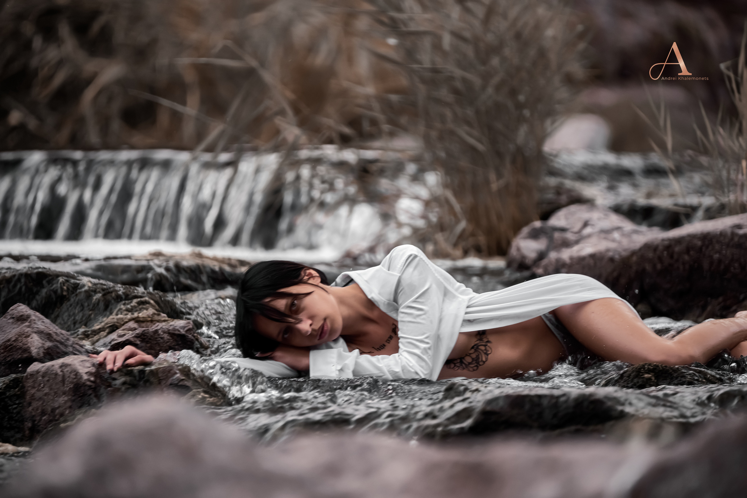 Обнаженная девушка в мокрой белой прозрачной рубахе лежит на камнях у водопада.
