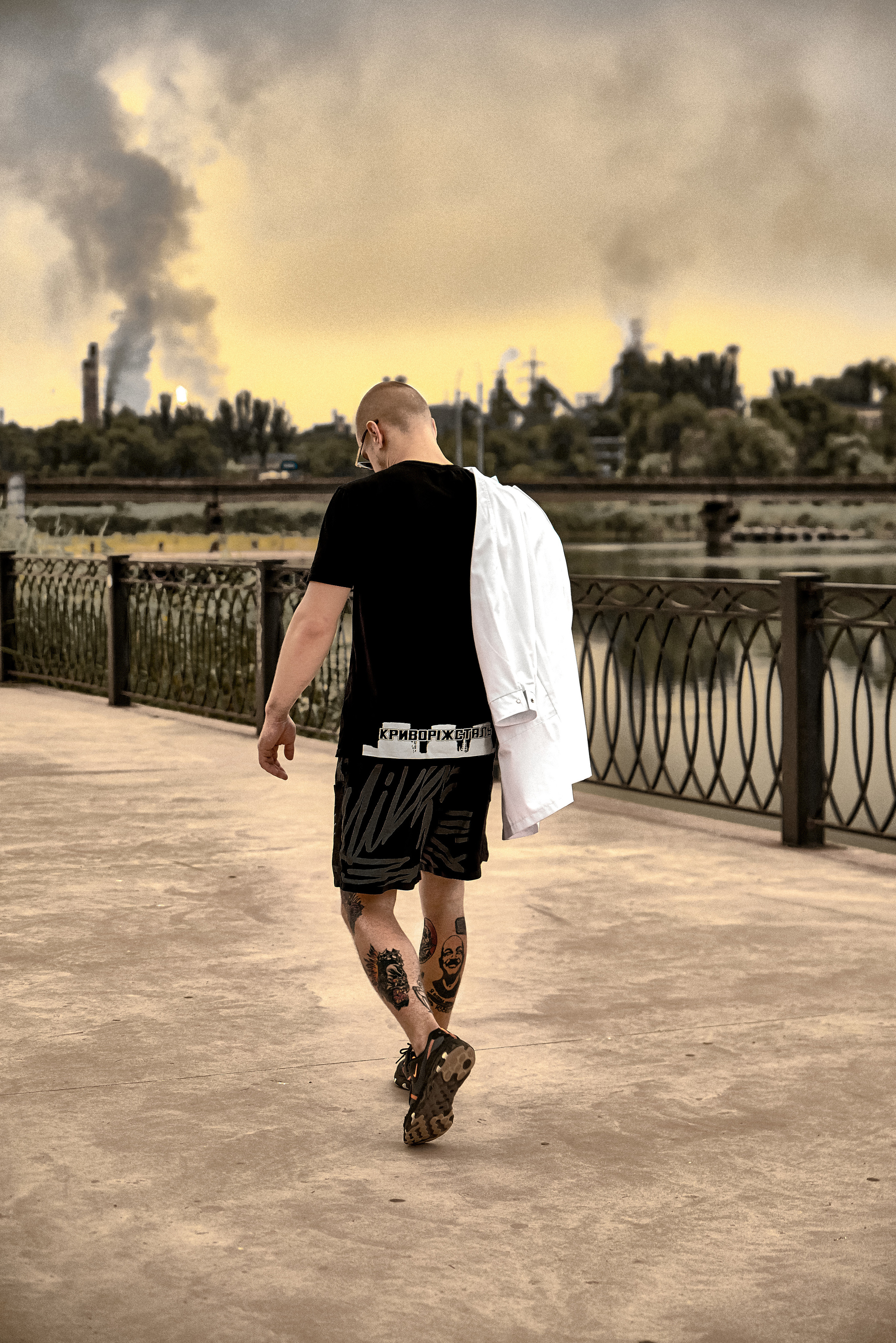 Спортивный парень с тату и белым халатом на плече идет по набережной на фоне черного дыма завода.