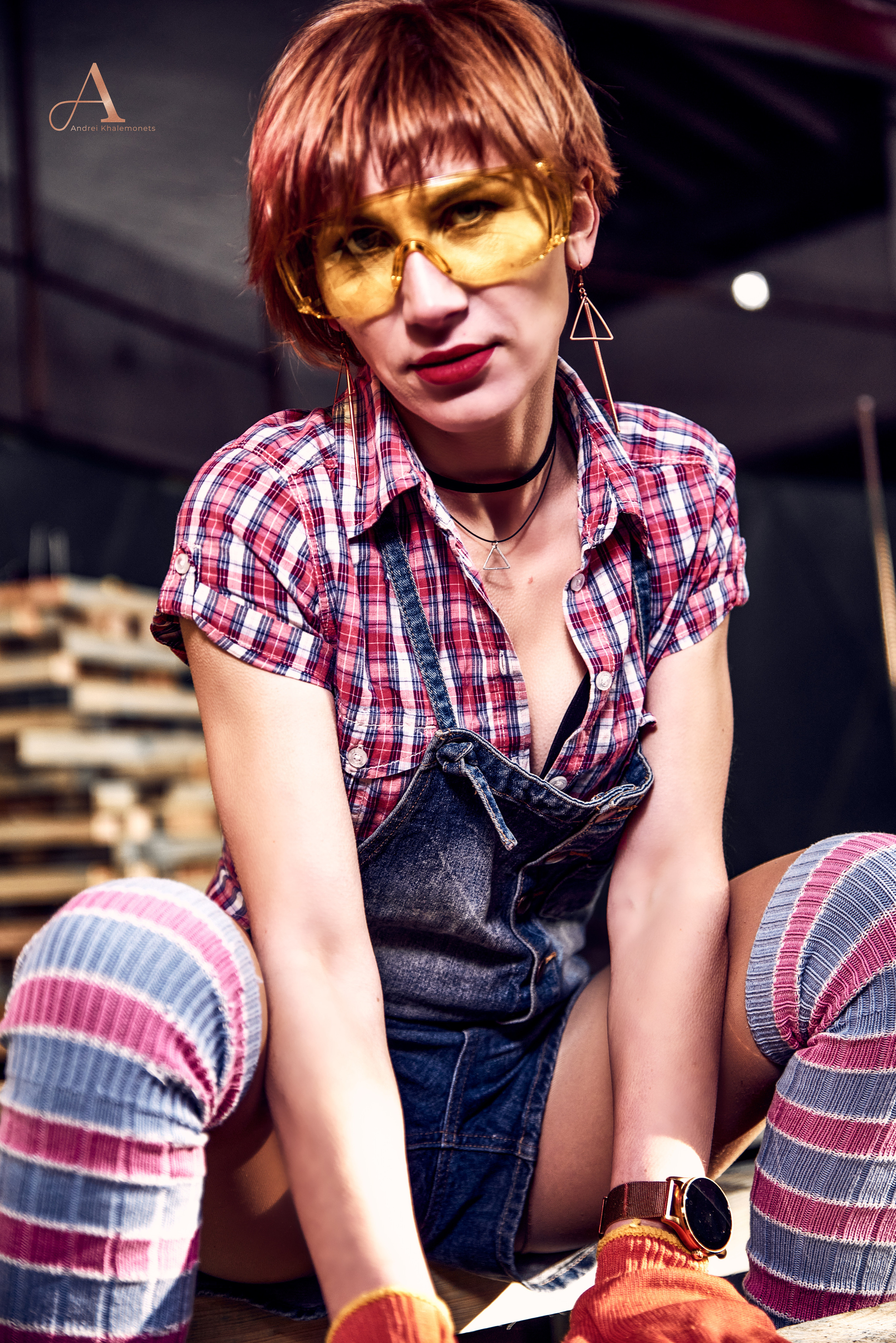 Девушка в защитных очках позирует в джинсовом комбинезоне и клетчатой рубашке.