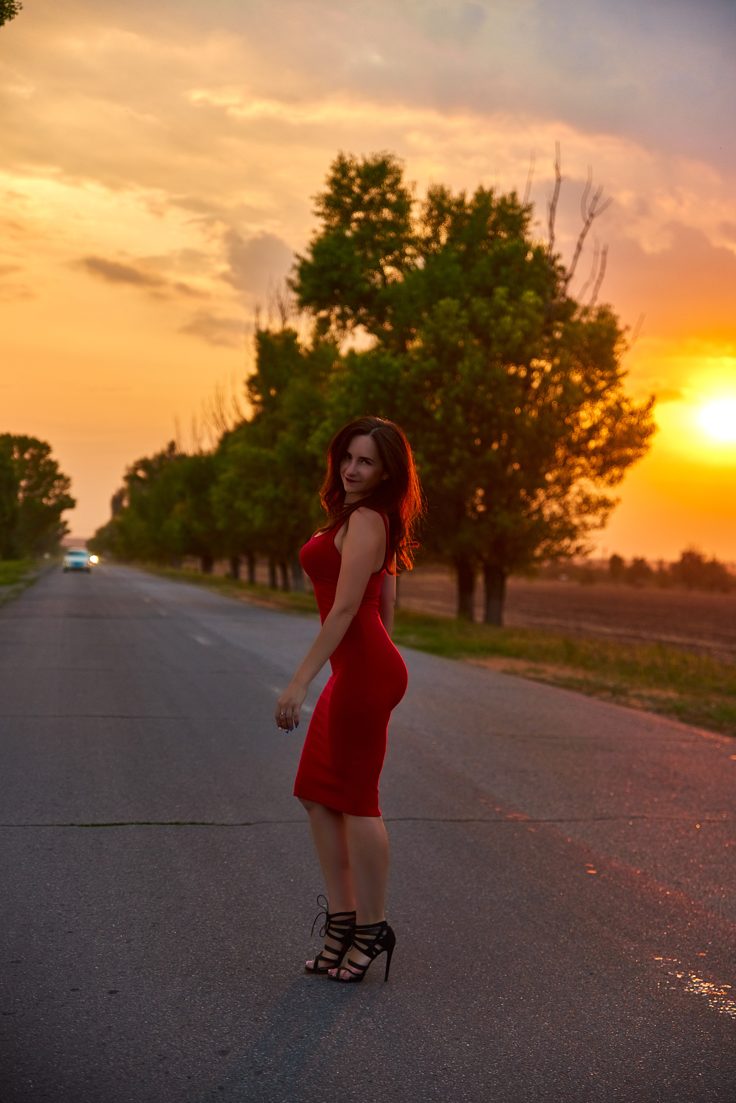 Девушка в красном платье на каблуках гуляет по автобану на закате.