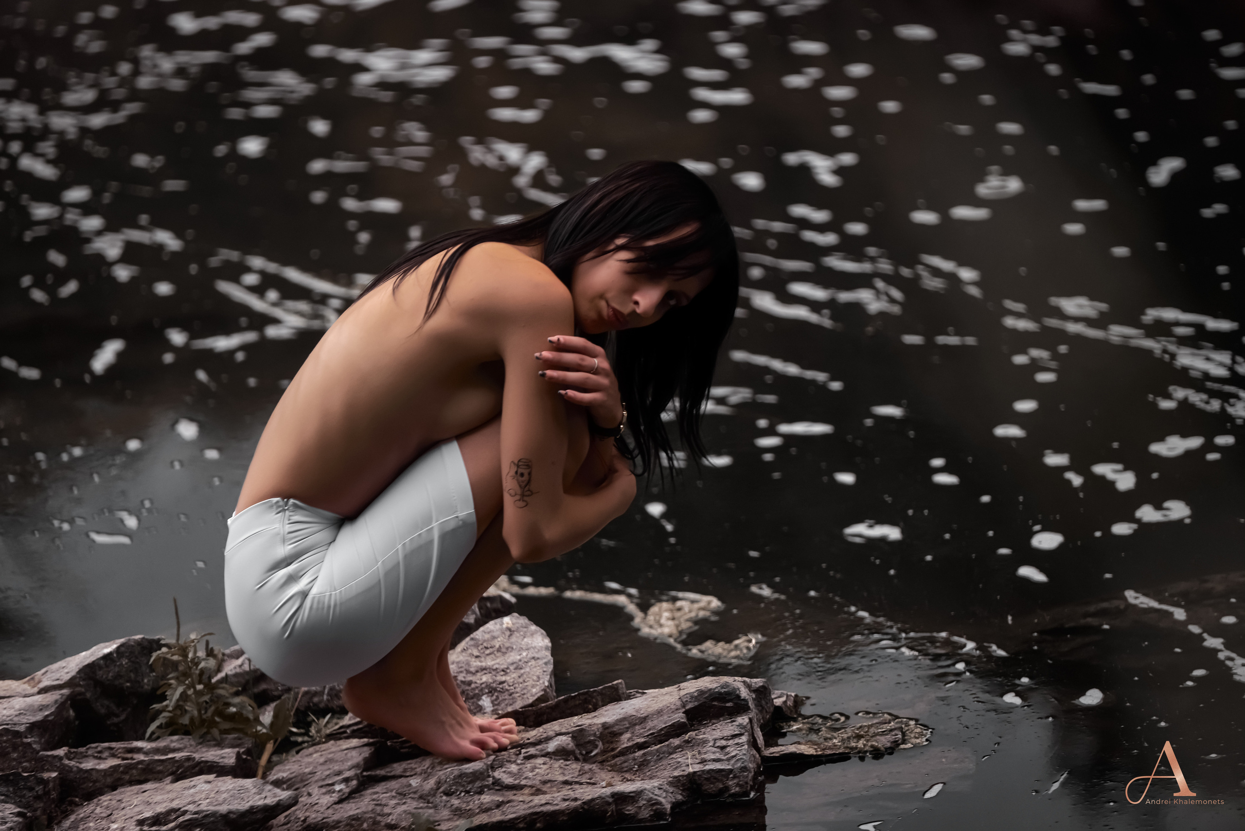 Девушка топлес в узкой белой юбке сидя обнимает себя за колени у водоема.